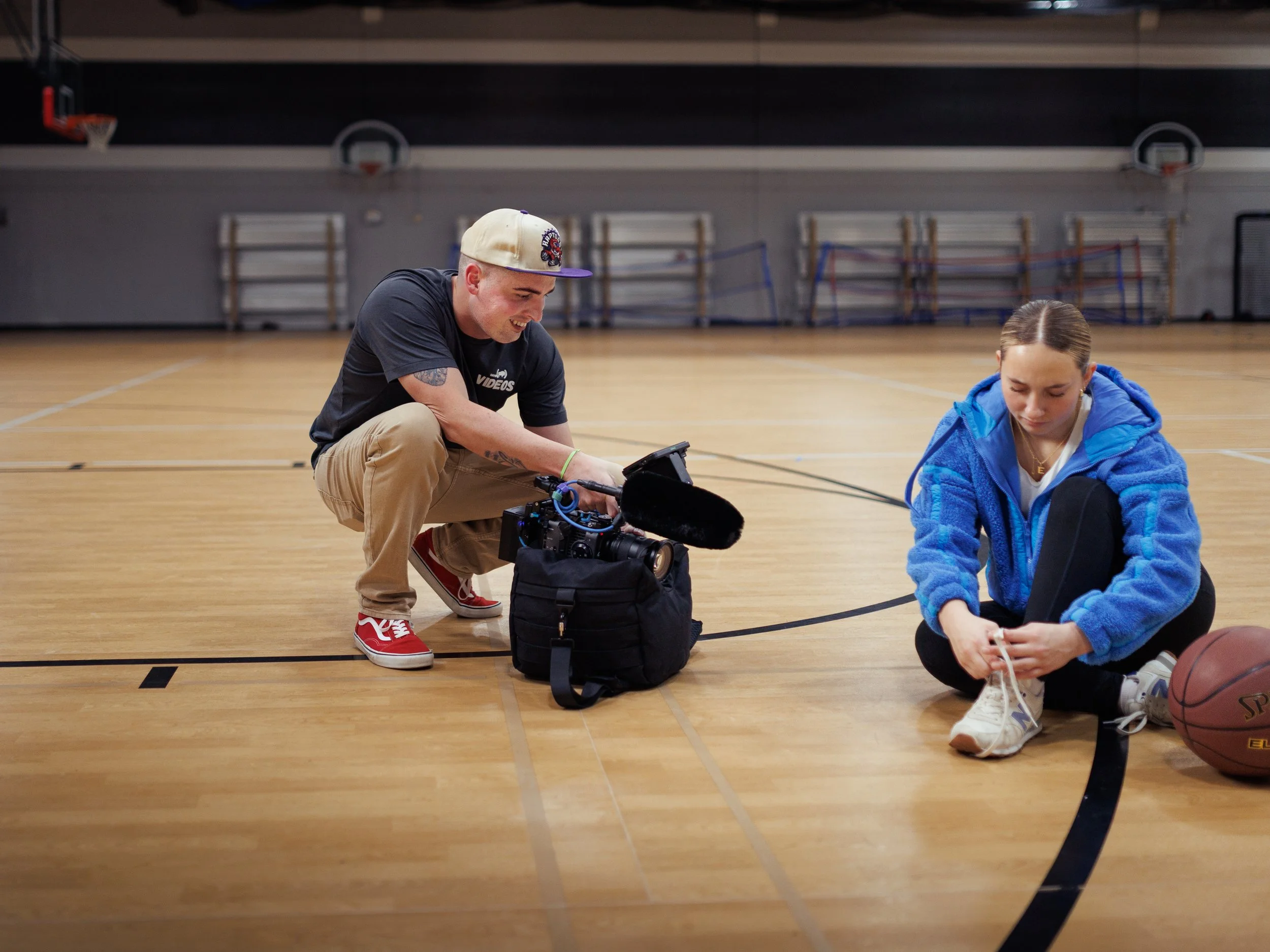 Behind-the-scenes in a gym: a videographer from Gavin (Bison) Videos crouches by a camera bag adjusting a rig with a shotgun mic while a young athlete in a blue fleece ties her shoe; a basketball and hoops are visible.