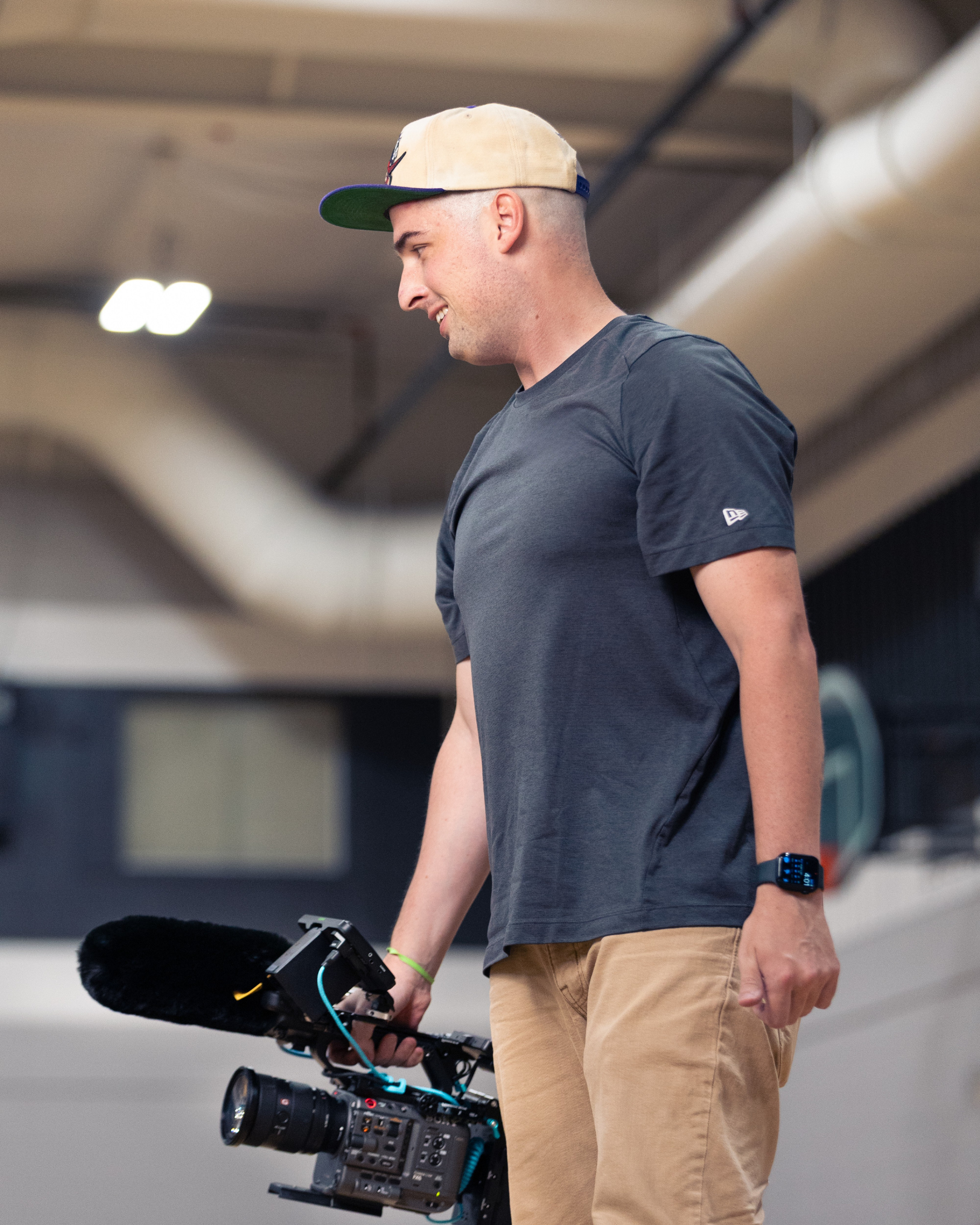 Gavin Gill, owner of Gavin (Bison) Media, wearing a beige baseball cap, a black t-shirt, tan pants, and a smartwatch holding a professional video camera in an indoor setting laughing with a actor/actress on set before the scene was filmed.