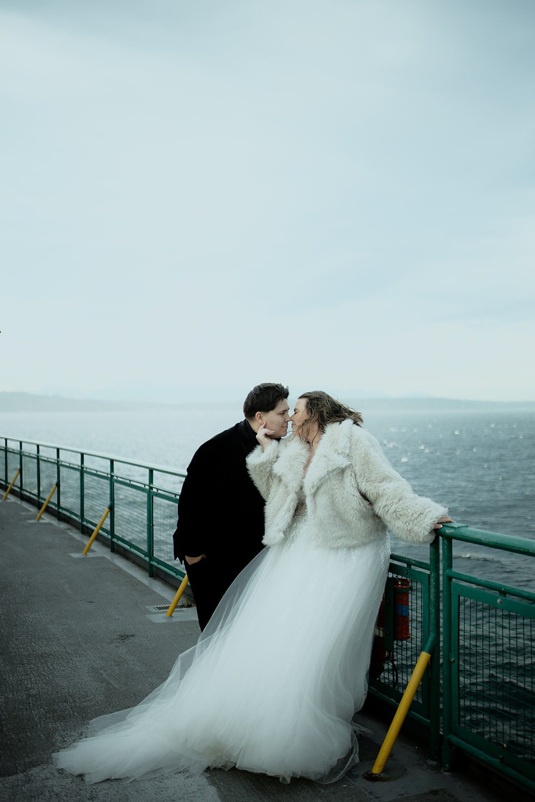 A couple, dressed in wedding attire, is leaning close to each other on a bridge by the water.