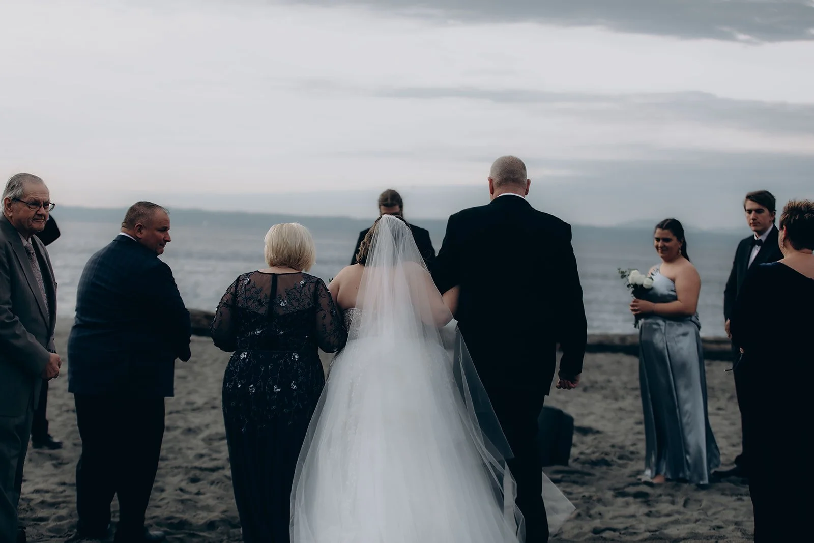 Wedding ceremony on the beach with the bride in a white gown and veil, surrounded by family and friends, with a cloudy sky and ocean in the background.