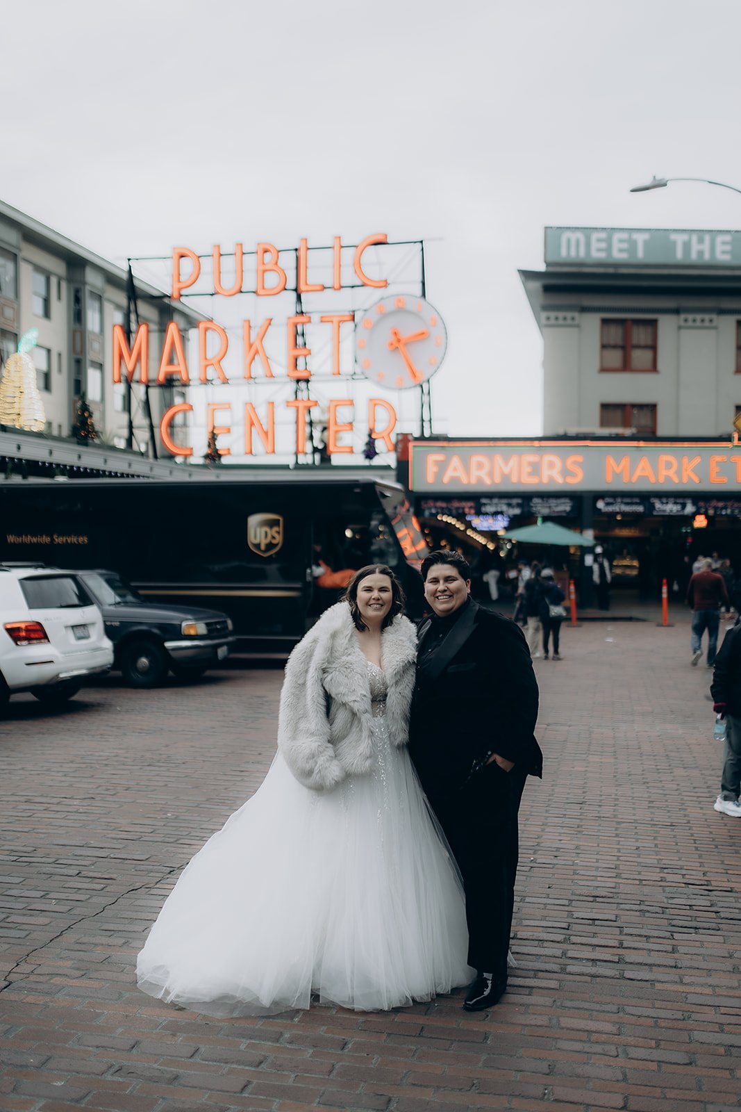 A couple dressed in wedding attire standing in front of a neon sign for Pike's Public Market Place. The bride is wearing a white gown and a fur coat, and the other bride is in a black suit.