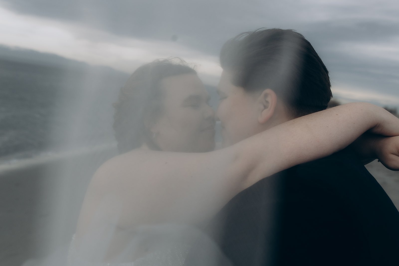 A couple embracing closely on a beach, with the woman's arm wrapped around the other woman's neck, both with eyes closed, under a cloudy sky.
