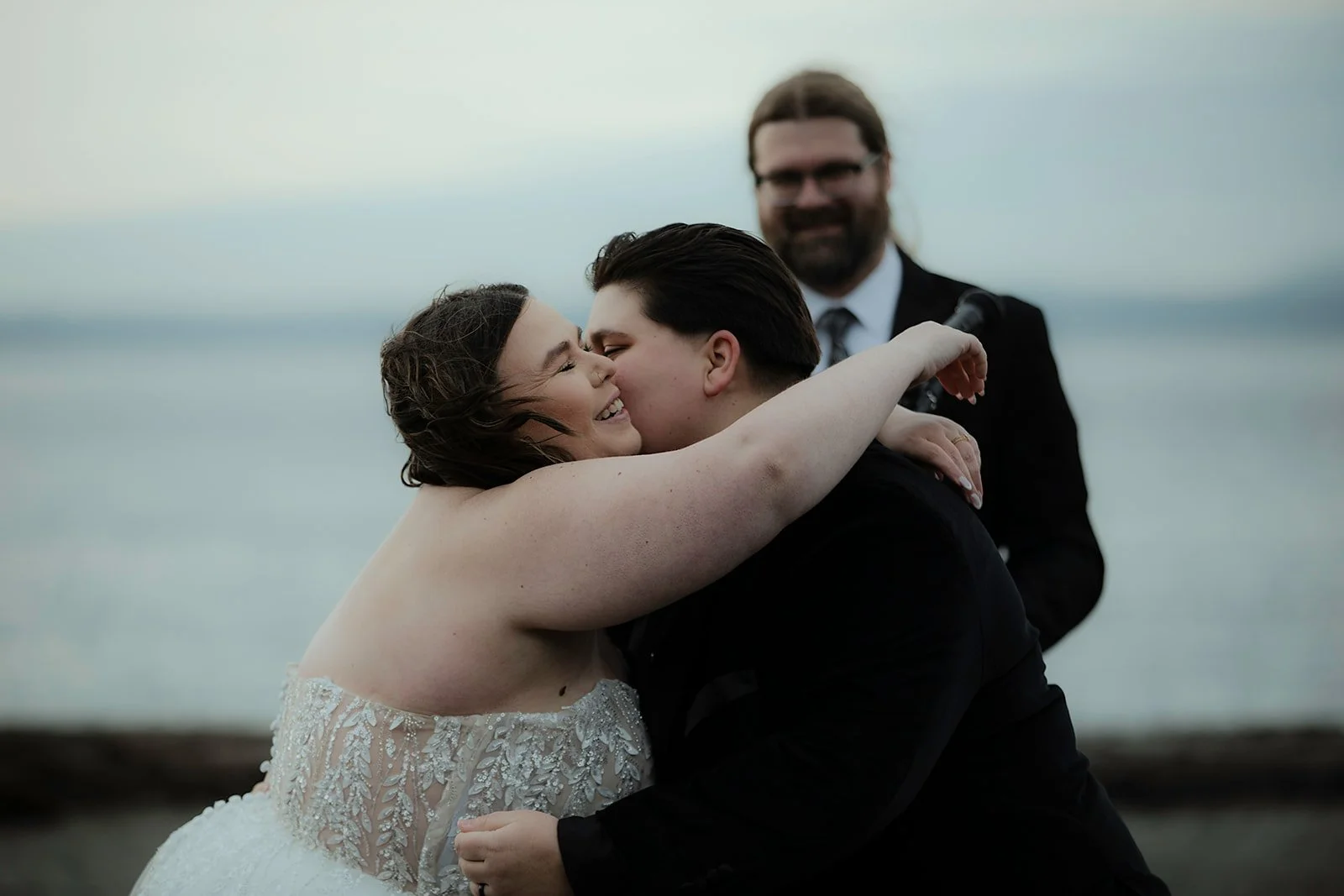A newlywed couple shares a kiss and hug, with a smiling officiant or witness in the background, near a body of water during sunset or dusk.