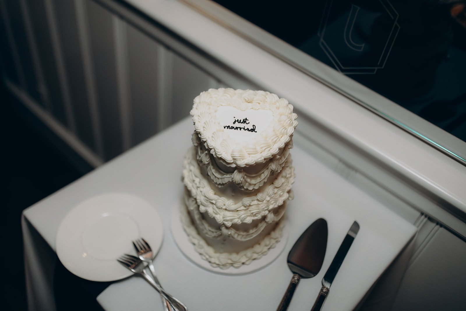 A three-tiered heart-shaped wedding cake decorated with white frosting that says 'just married' in black script, placed on a white table with serving utensils and a small empty plate.