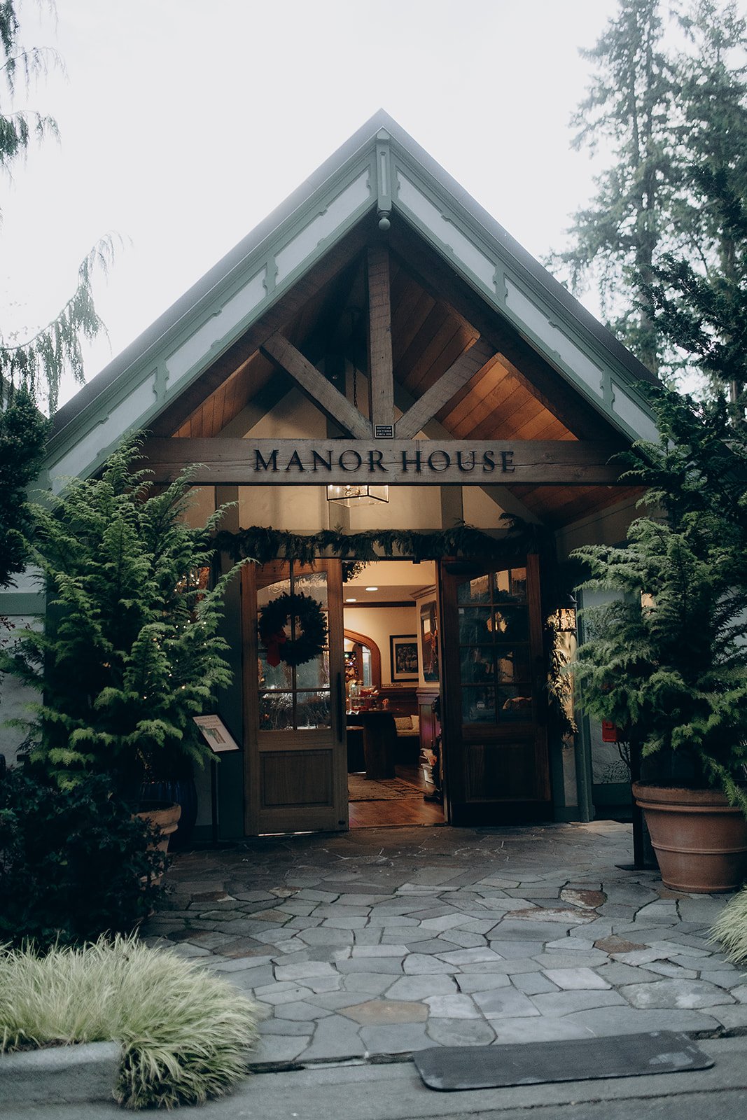 Exterior of Manor House with open wooden doors, potted plants, and holiday decorations inside.