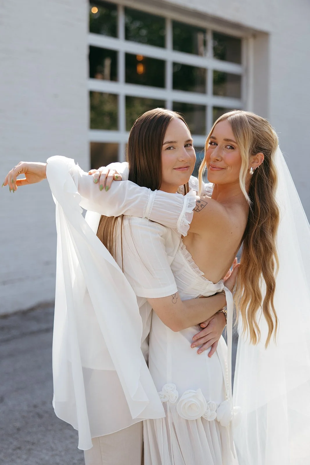Two women embracing outdoors, one dressed in a wedding gown and the other in a white suit, in front of a modern building with large windows.