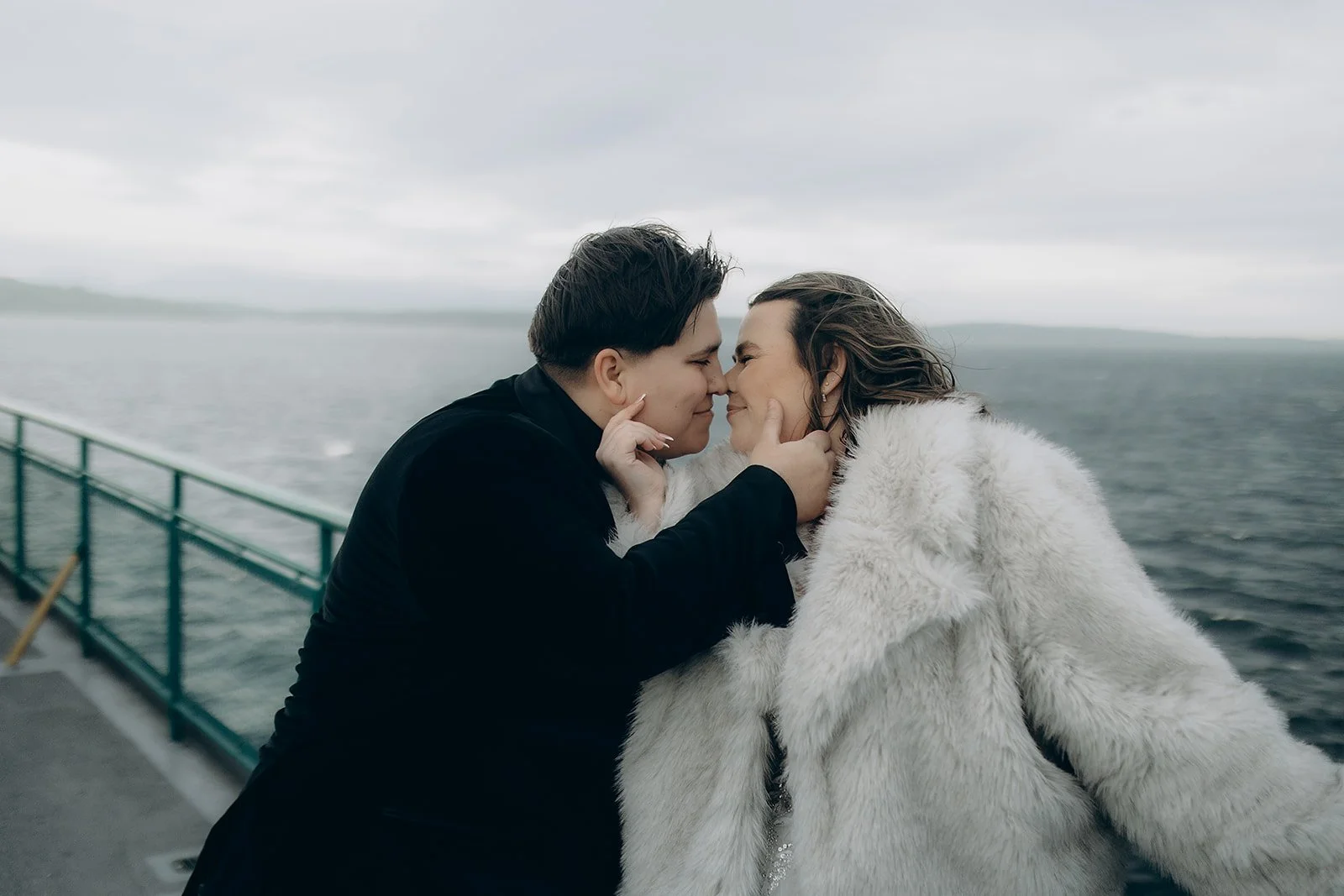 Two brides on a ferry, about to kiss, with water and an overcast sky in the background.