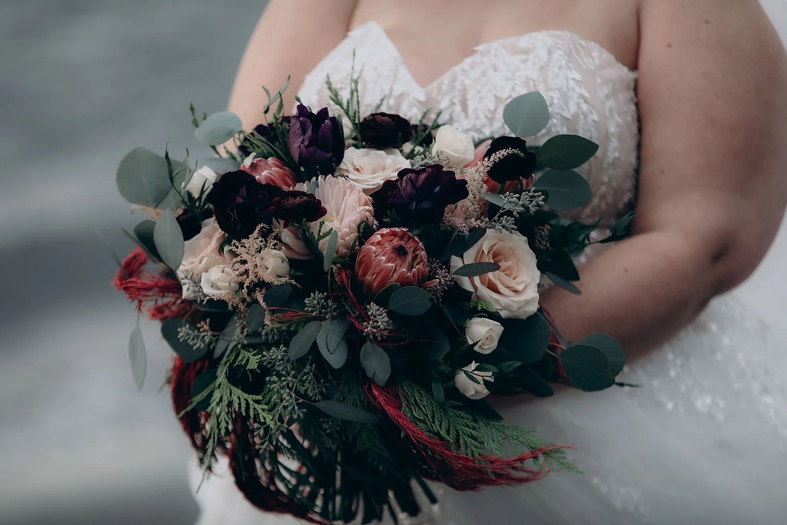 A bride in a white wedding dress holding a bouquet of dark purple, blush pink, and cream flowers with greenery.