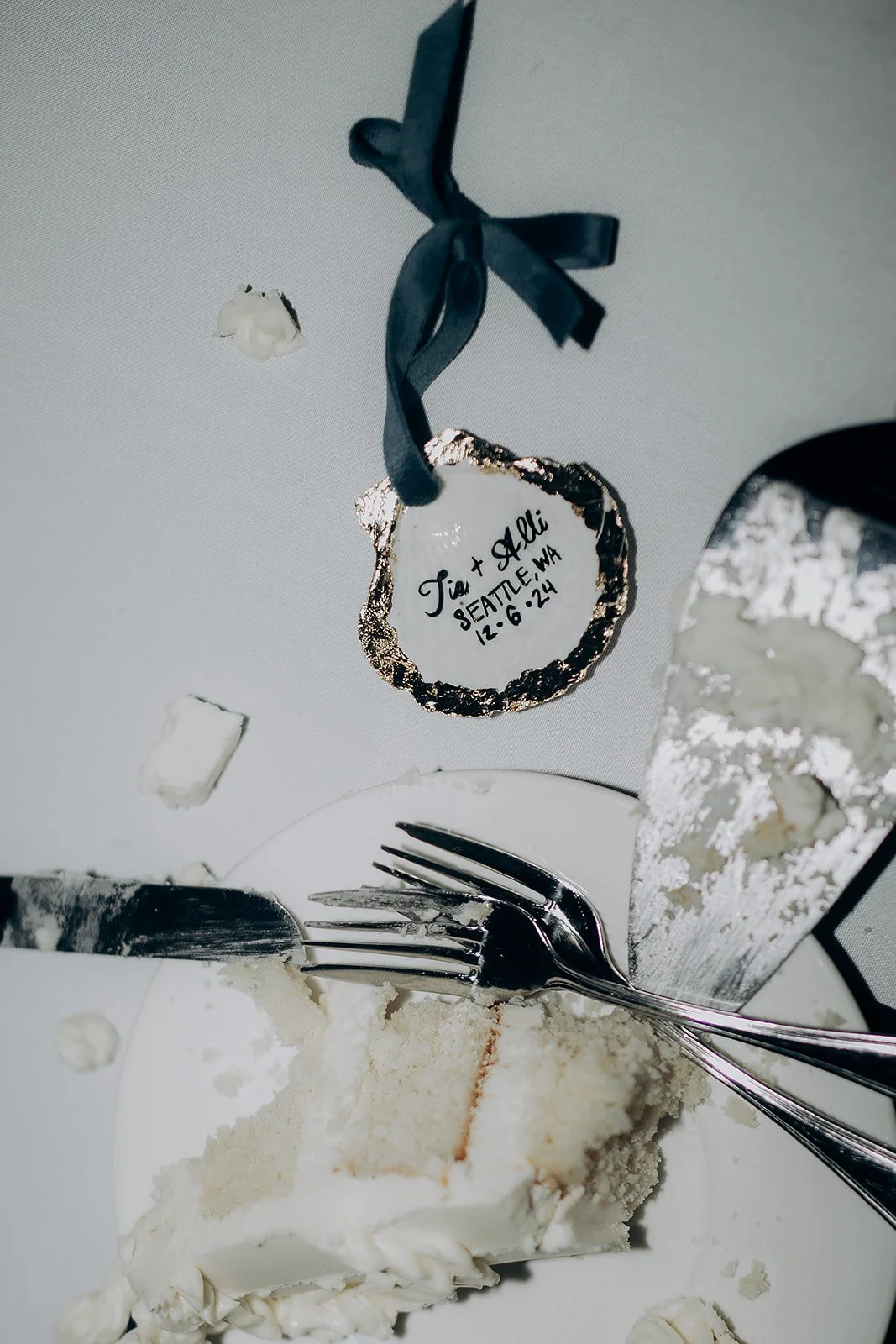 Wedding cake with cake knife and server on a table, a small decorative piece with a black ribbon and an inscription note indicating the wedding of Tia and Alli in Seattle, Washington on December 6, 2024, with some crumbs on the table.
