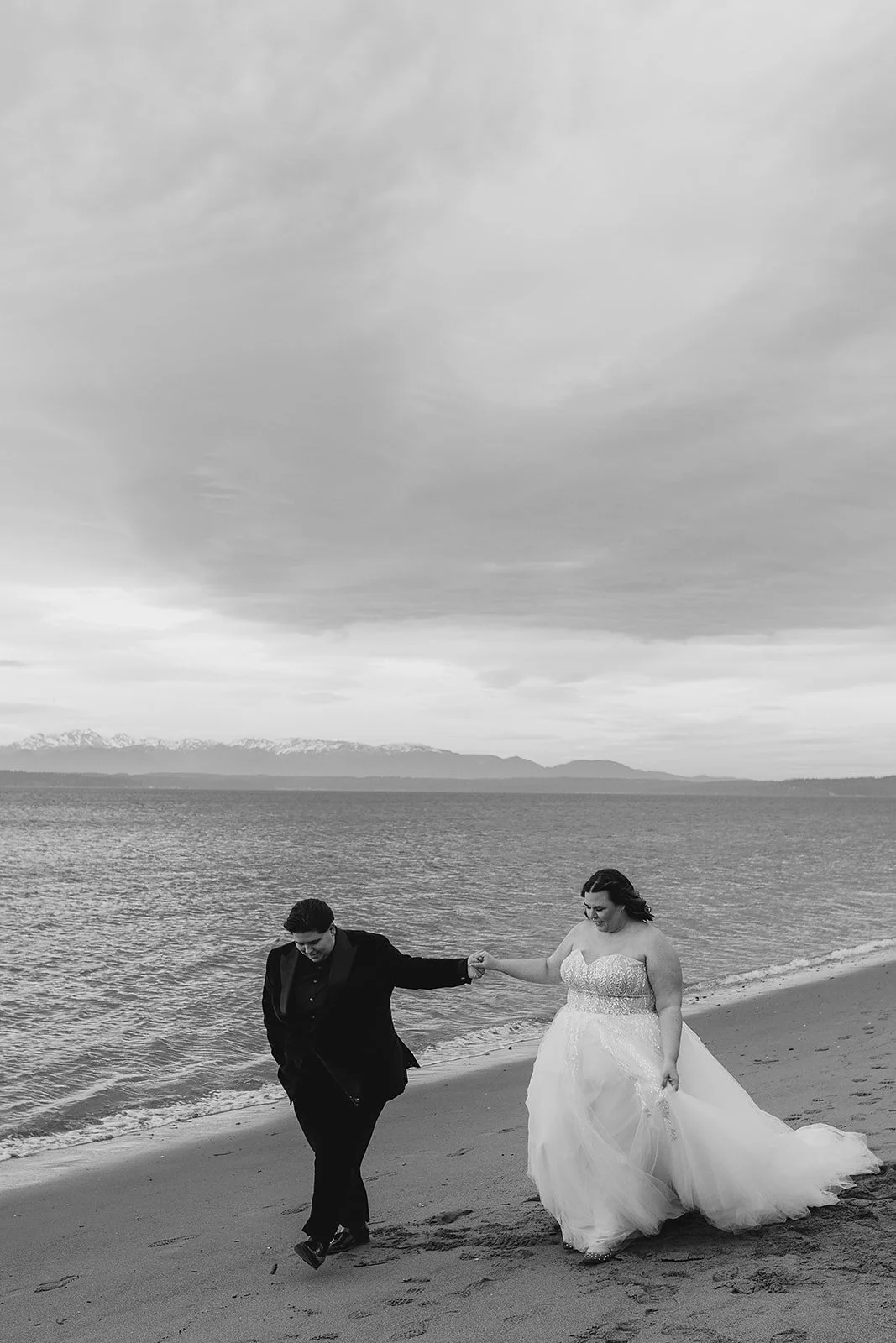A black-and-white photo of a couple in wedding attire walking hand in hand along a beach, with mountains in the background.