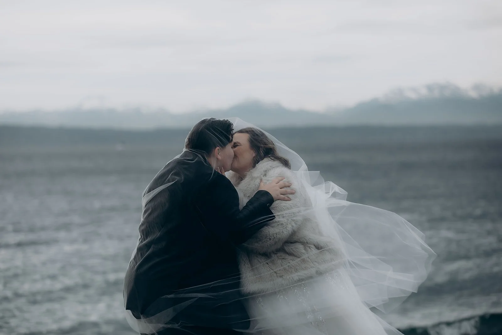 two women kiss on a beach with mountains in the background