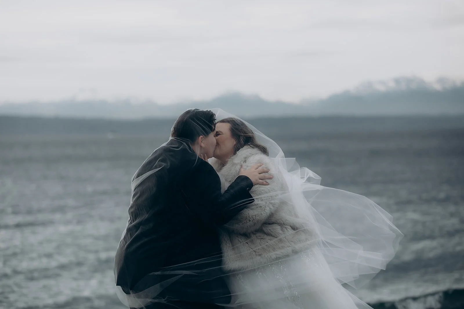 Two women in wedding attire sharing a kiss by the water on a cloudy day, one wearing a black jacket and the other a white wedding dress with a veil.