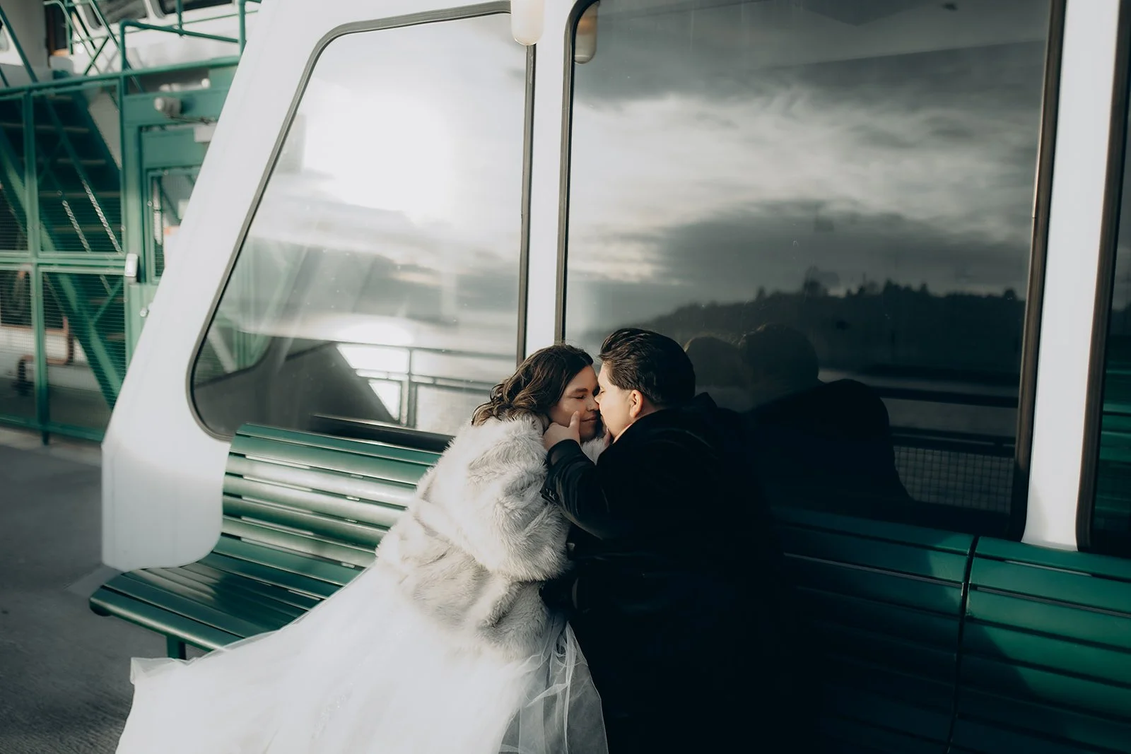 A couple sitting on a green bench inside a modern transportation station, sharing an intimate moment with their foreheads touching and eyes closed. 