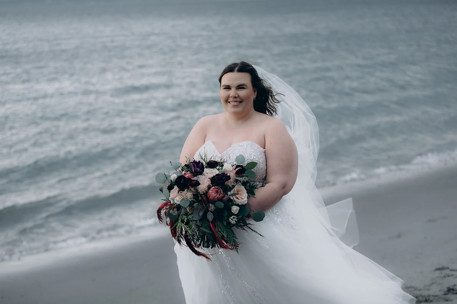 Bridal portrait of a smiling woman in a wedding gown holding a bouquet of flowers, on a beach with the ocean in the background.