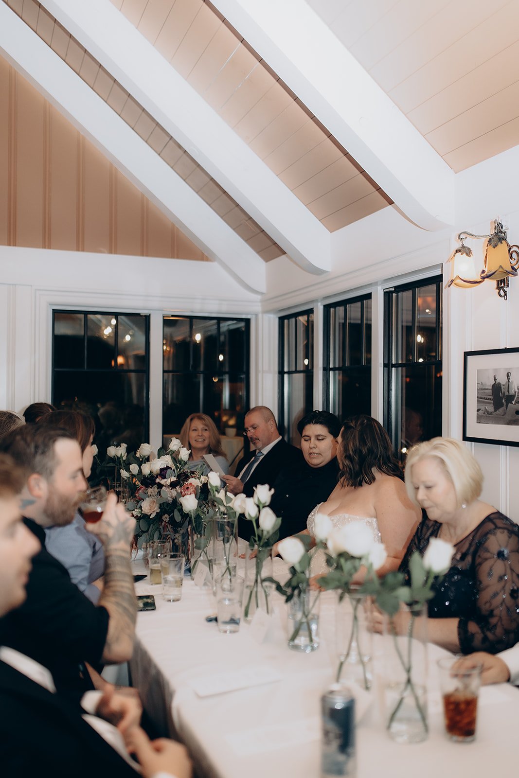 A group of people sitting at a long dining table decorated with white roses in clear vases, inside a brightly lit room with wood-paneled ceiling and large windows.