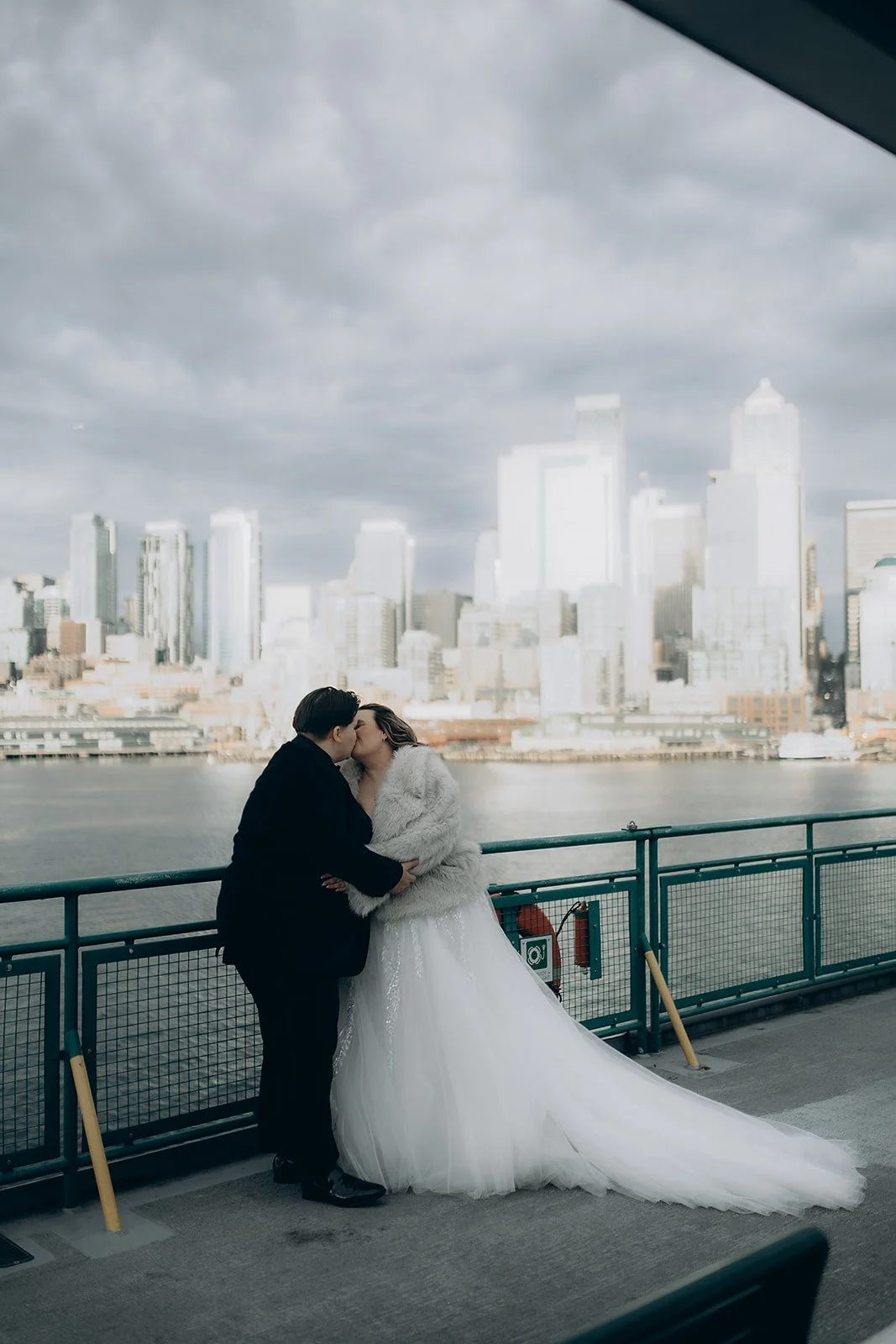 A couple in wedding attire sharing a kiss on a boat deck overlooking a city skyline.