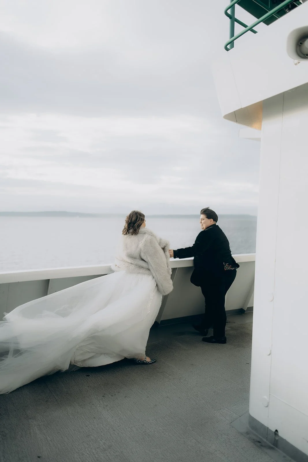 A bride in a wedding dress and faux fur coat stands on a ship's deck, holding hands with a woman in black, as they look out over the water on a cloudy day.