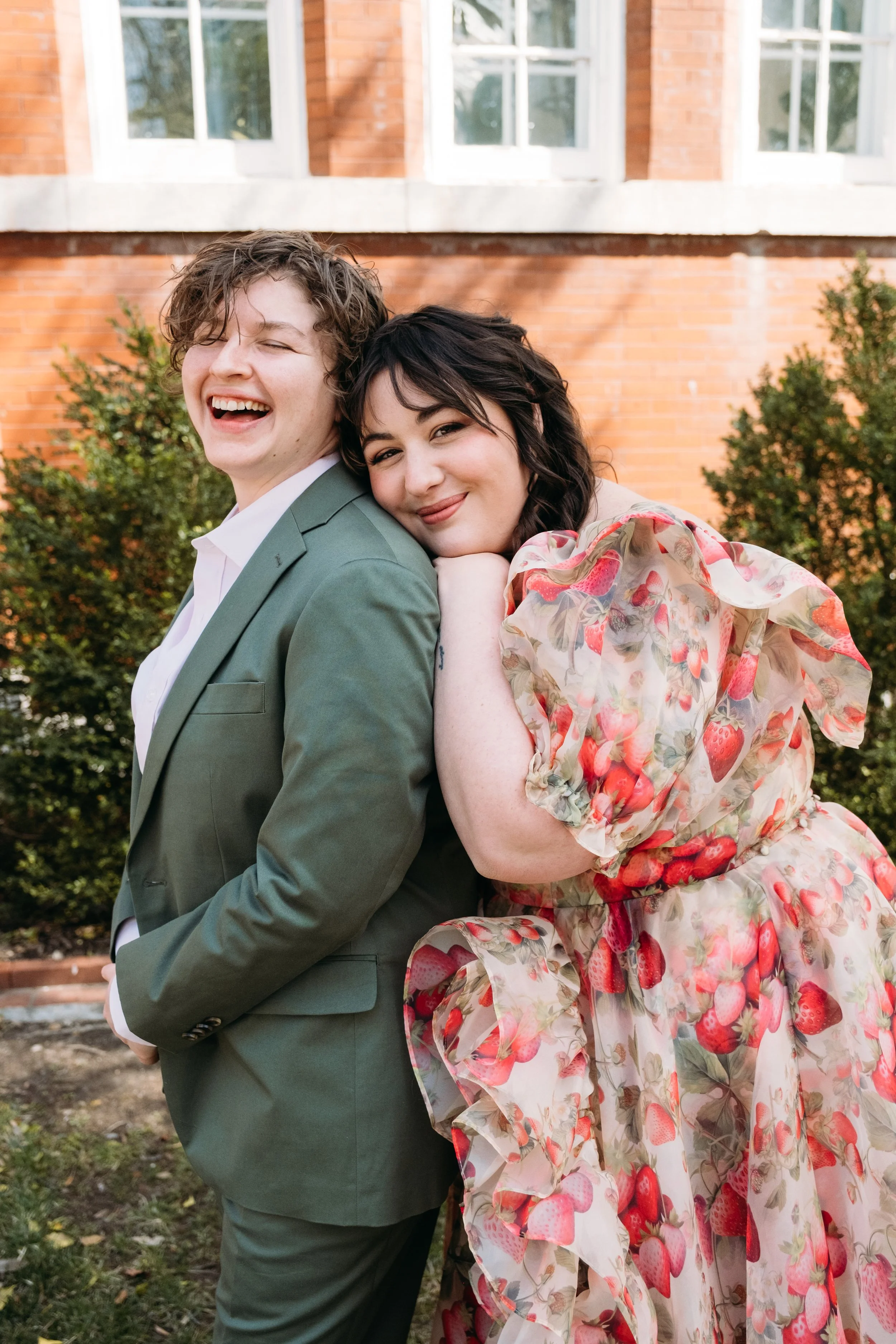 A person in a green suit and a woman in a floral dress are standing close together outdoors in front of a brick building with large windows. 