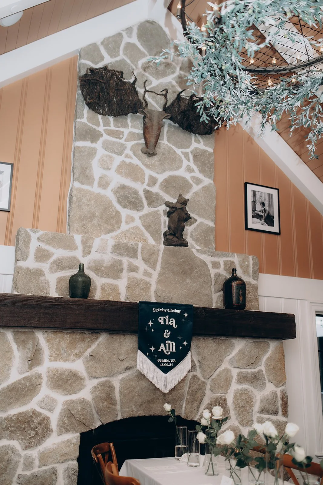 A rustic stone fireplace with a mount of a deer head with antlers, surrounded by a bear sculpture, vases, and a wedding banner that reads 'Tia & Ali, Seattle, WA, 12.06.24.' There are framed photographs on the walls and a table set with white flowers