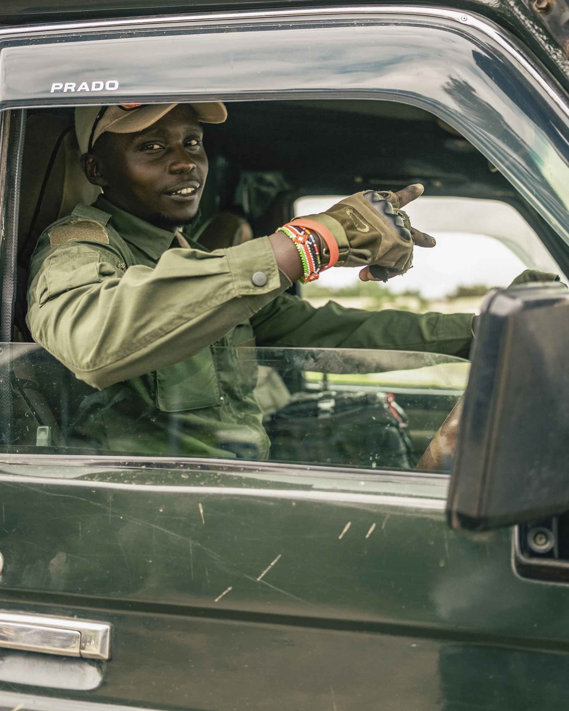 A man sitting in a vintage green car with a "PRADO" emblem on the side, pointing outside the car window, wearing a beige cap, military-style shirt, and colorful beaded bracelets.