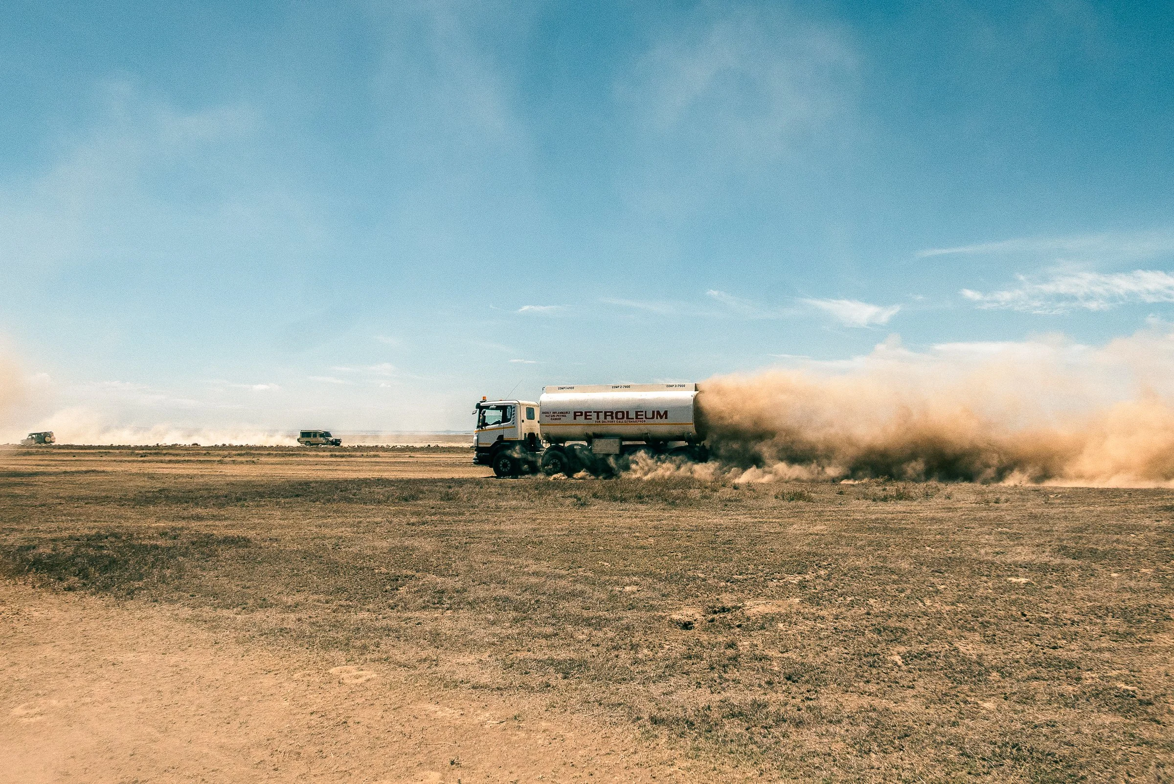 A white petroleum tanker truck driving on a dusty, open plain with dust clouds behind it, under a mostly clear sky.