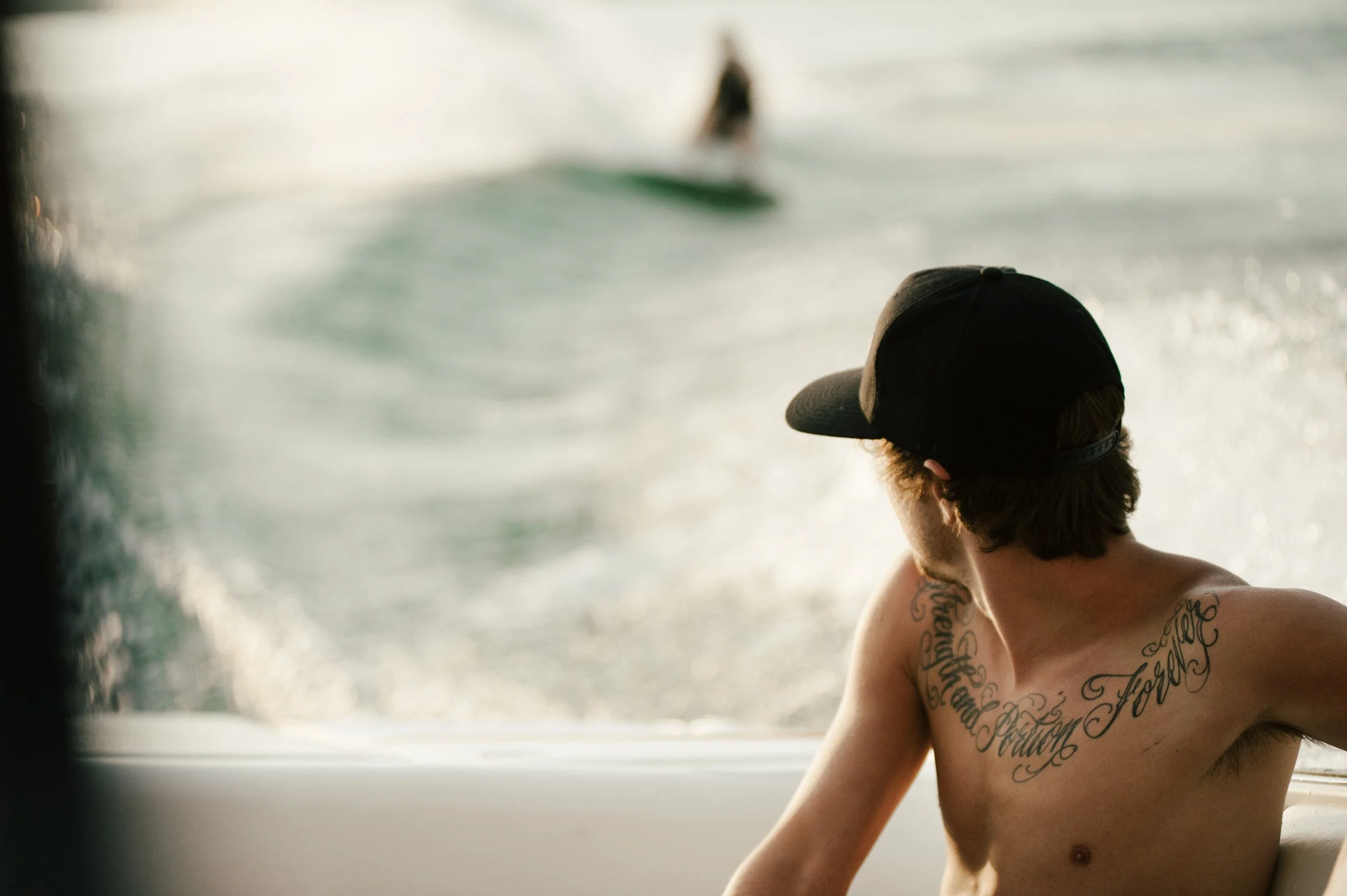 A young man with tattoos wearing a black cap, sitting shirtless near a window by the water, with a blurred boat in the background.