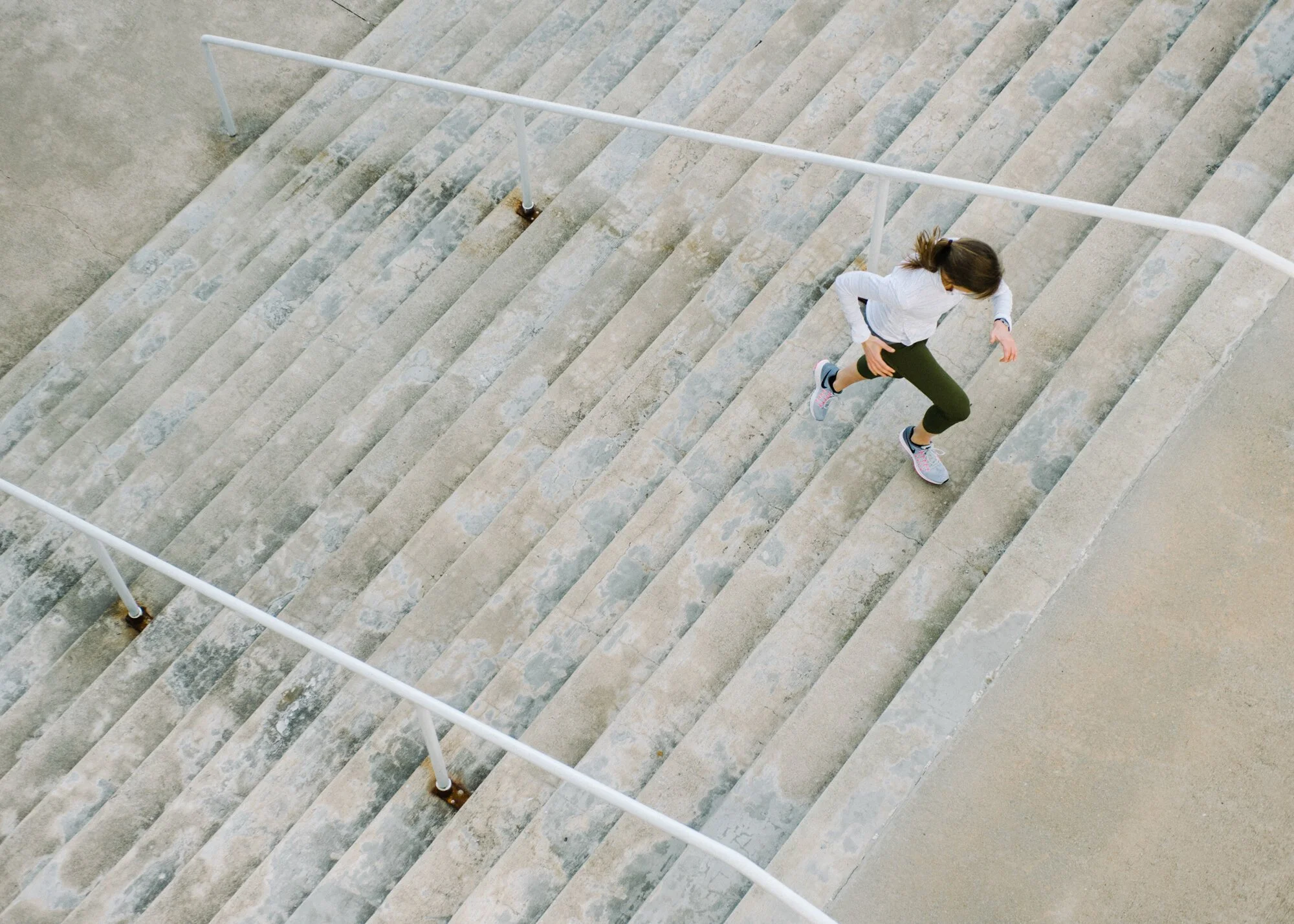 A woman in athletic clothing running up outdoor concrete stairs with metal handrails.