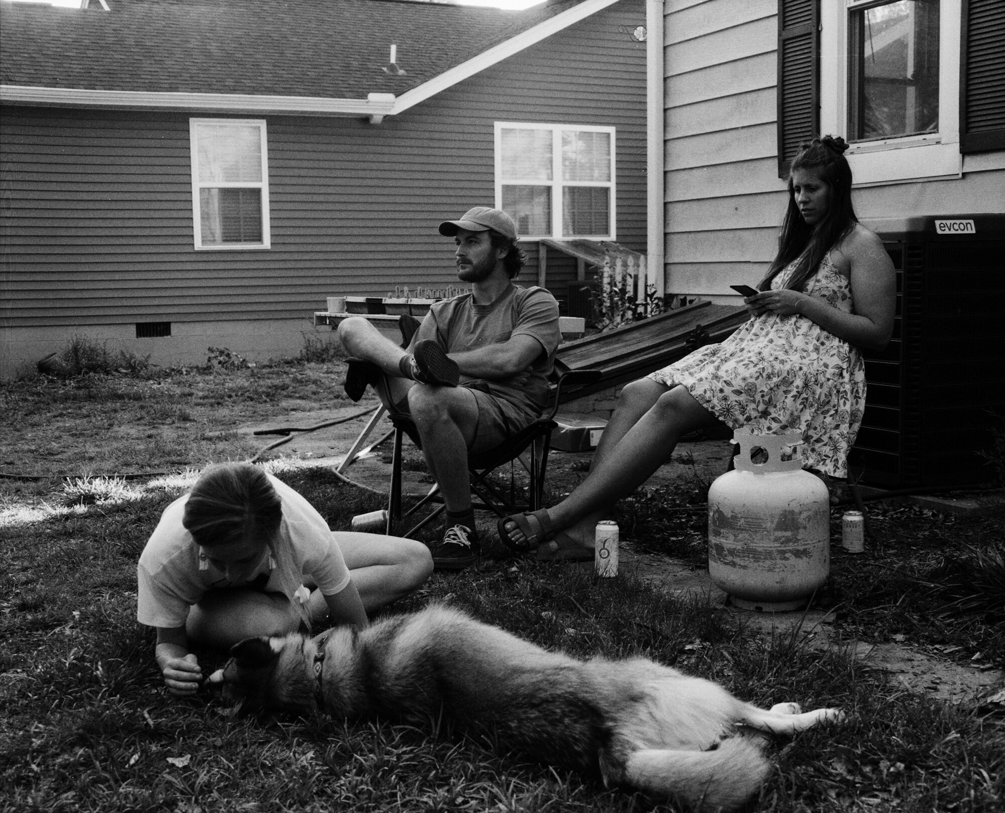 A young boy with glasses petting a dog lying on the grass, while a woman in a floral dress is sitting nearby and looking at her phone in the backyard of a house.