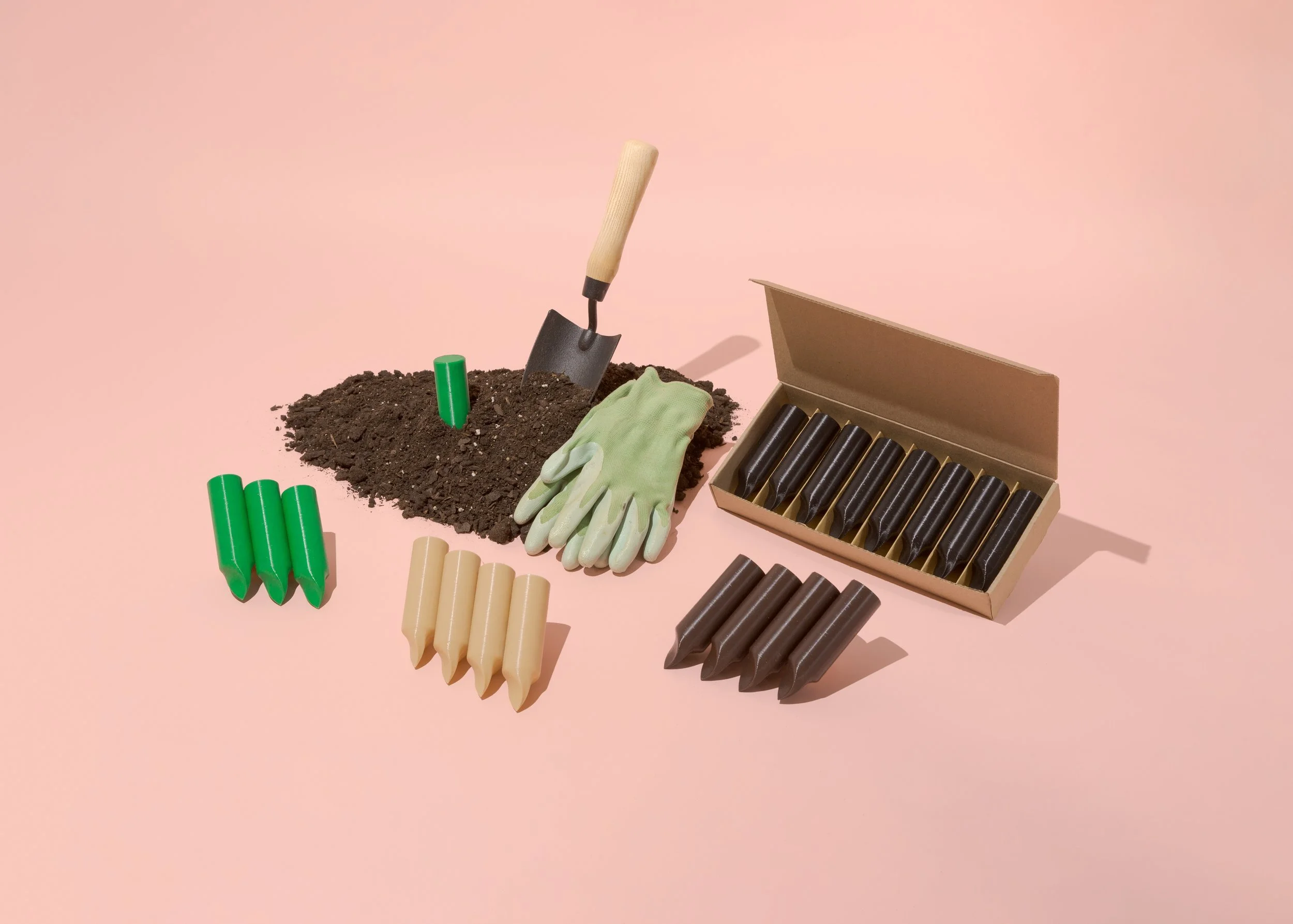 Farm supplies including a small gardening shovel, green and beige fertilizer spikes, a pair of work gloves, and a box of black seedling pots on a pink background.