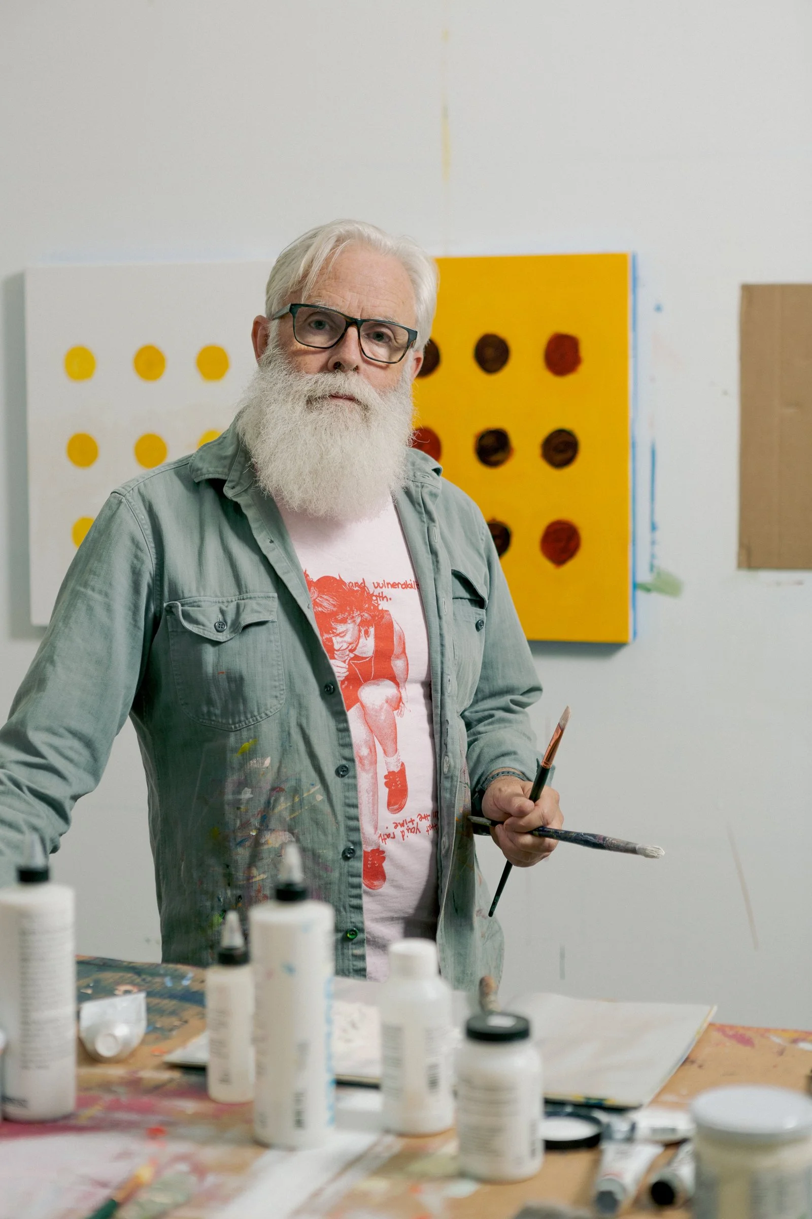 An elderly man with gray hair, glasses, and a white beard is standing in front of an art studio wall, holding a paintbrush, surrounded by bottles of paint on a table, with colorful paintings in the background.
