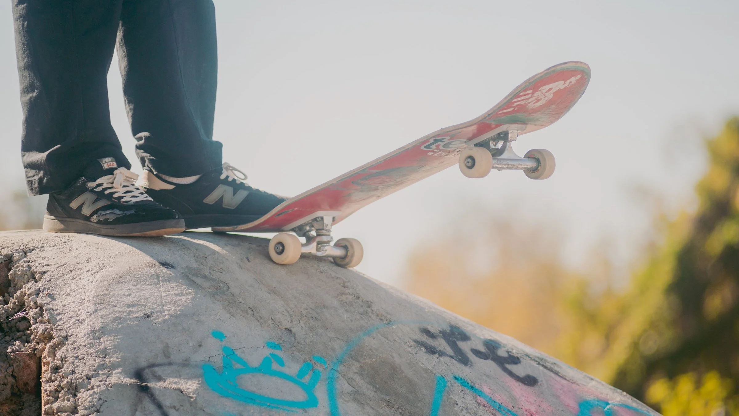 Close-up of a skateboarder's feet on a ramp, wearing black New Balance sneakers, with a skateboard positioned at an angle on the ramp in a skate park.