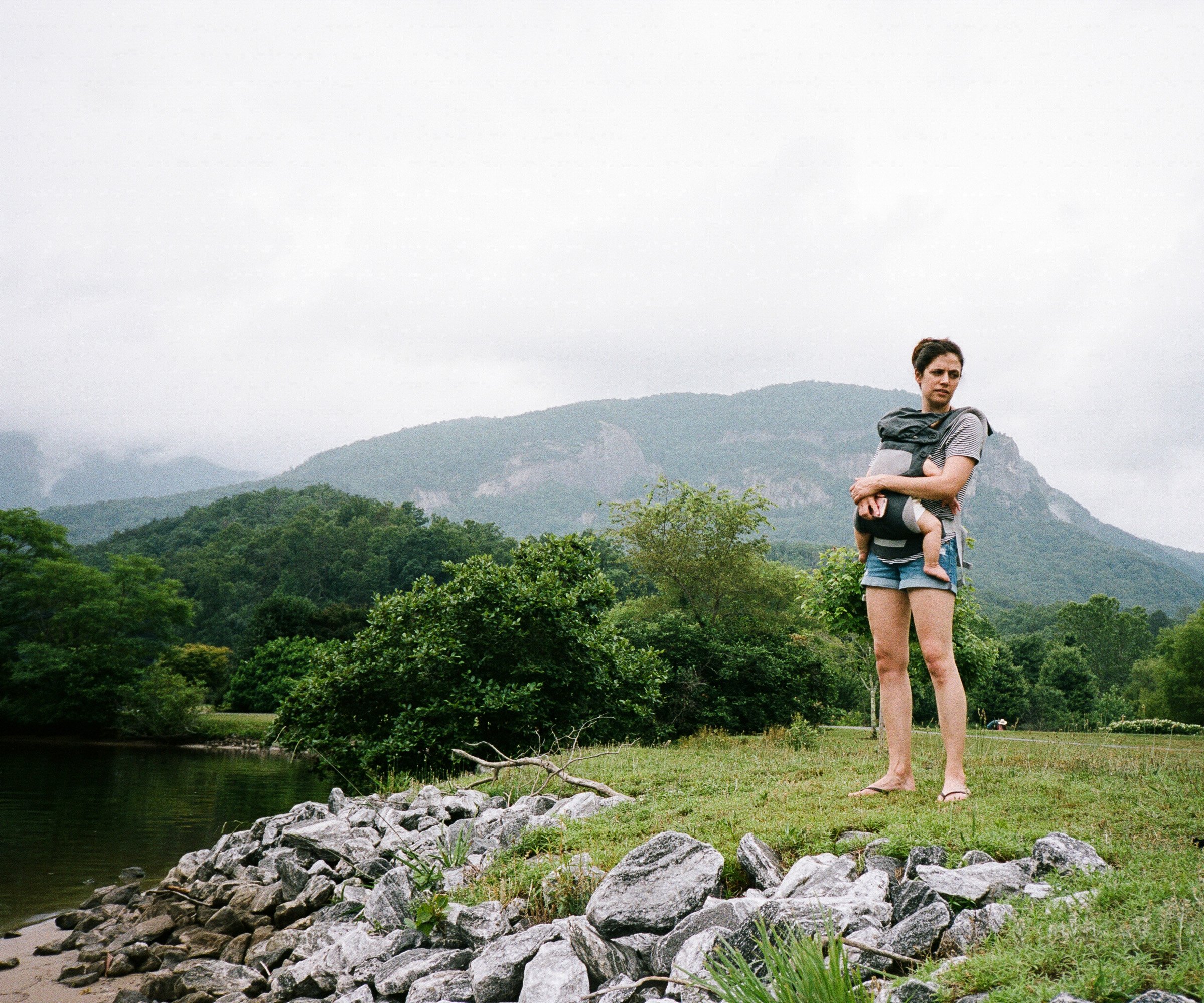 A woman is standing outdoors on a grassy area near a body of water, holding a child in her arms. She is dressed casually in shorts and a striped shirt. The landscape features lush green trees, mountains in the background, and an overcast sky.