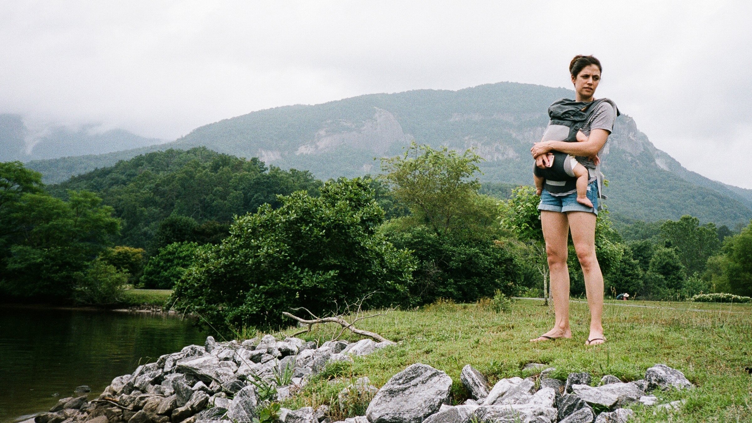 A woman with dark hair holding a child in a backpack carrier, standing on grass near a river, with lush green trees and mountains in the background on an overcast day.