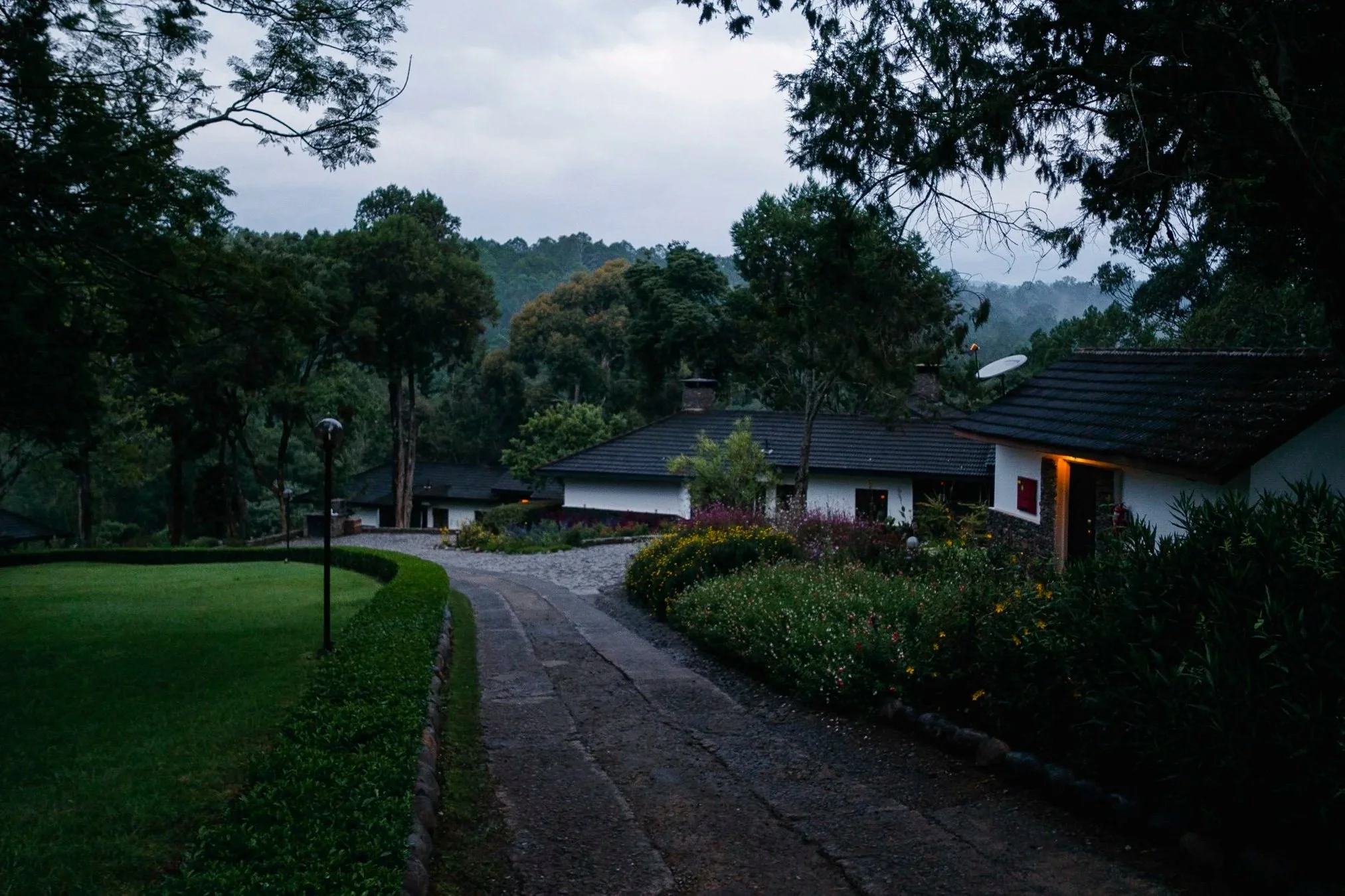 A residential area with houses, a winding paved pathway, lush gardens with colorful flowers, trees, and hills with dense forest in the background under an overcast sky.
