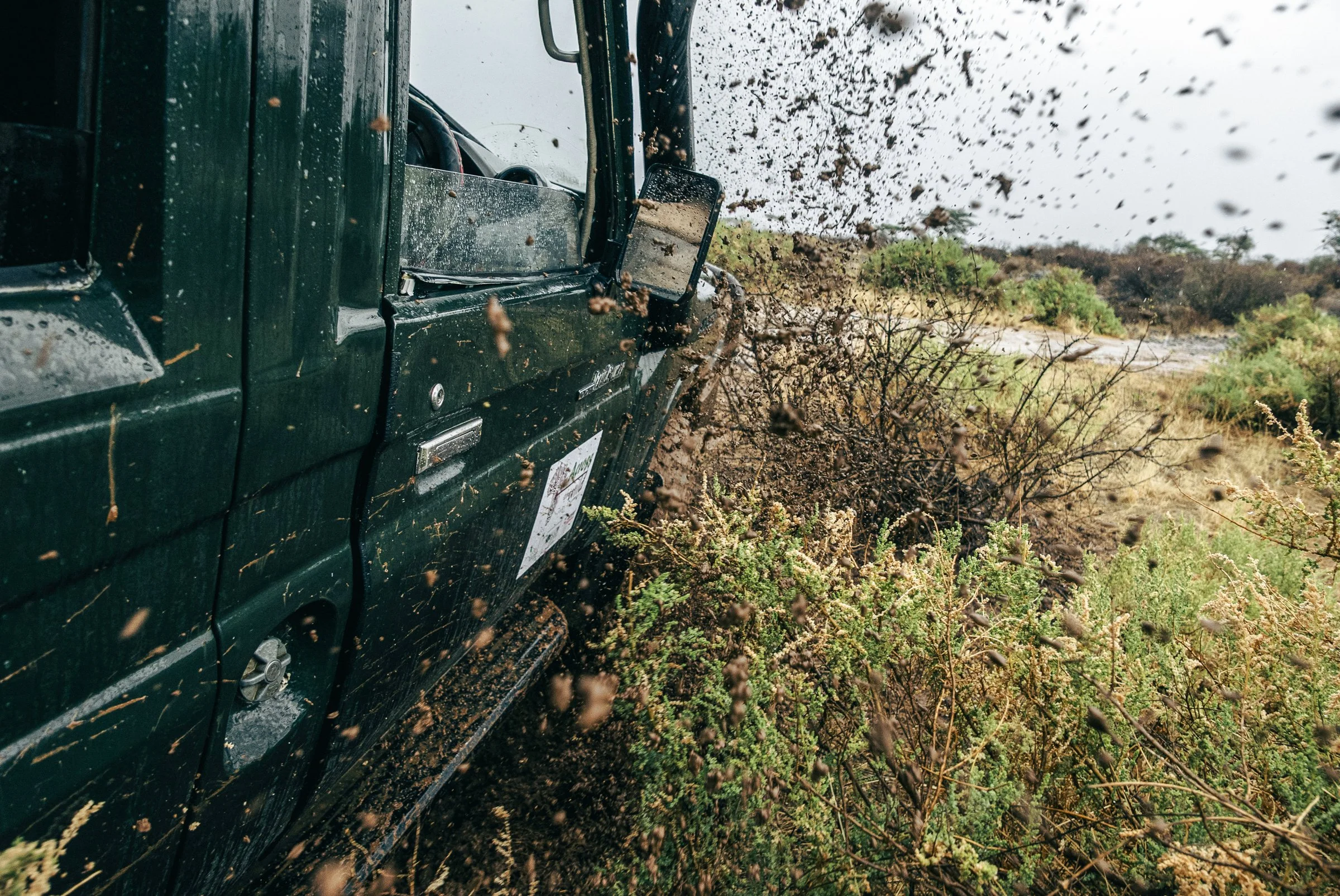 A green vehicle driving through muddy terrain with mud splashing up around it.