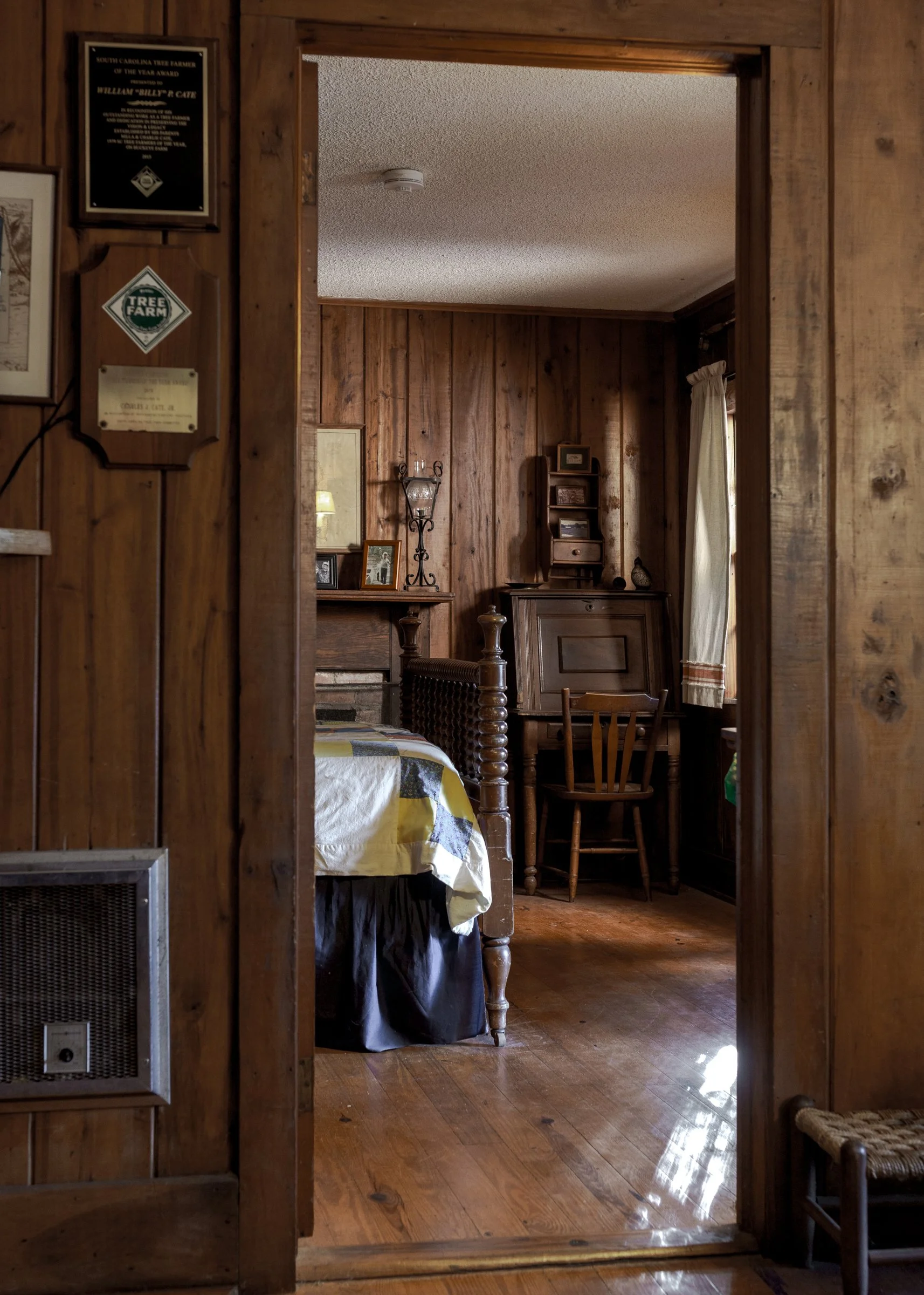 View of a rustic bedroom with wood-paneled walls and wooden furniture, including a bed with a quilt, a small bookshelf, a chair, and a window with curtains.