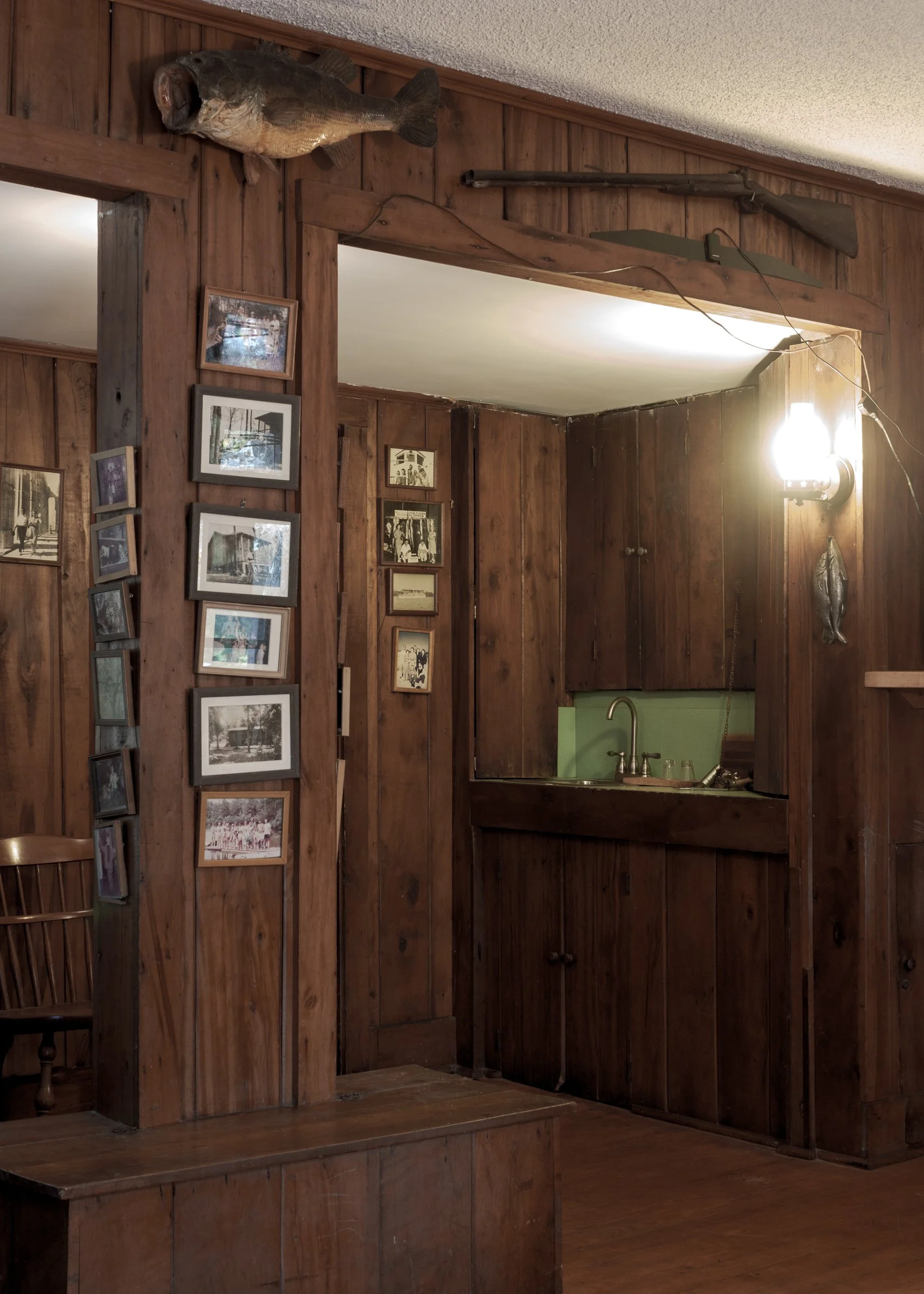 Interior of a room with wooden panel walls, displaying a mounted fish, a framed photo collage, a mounted gun, and a wall-mounted light fixture.