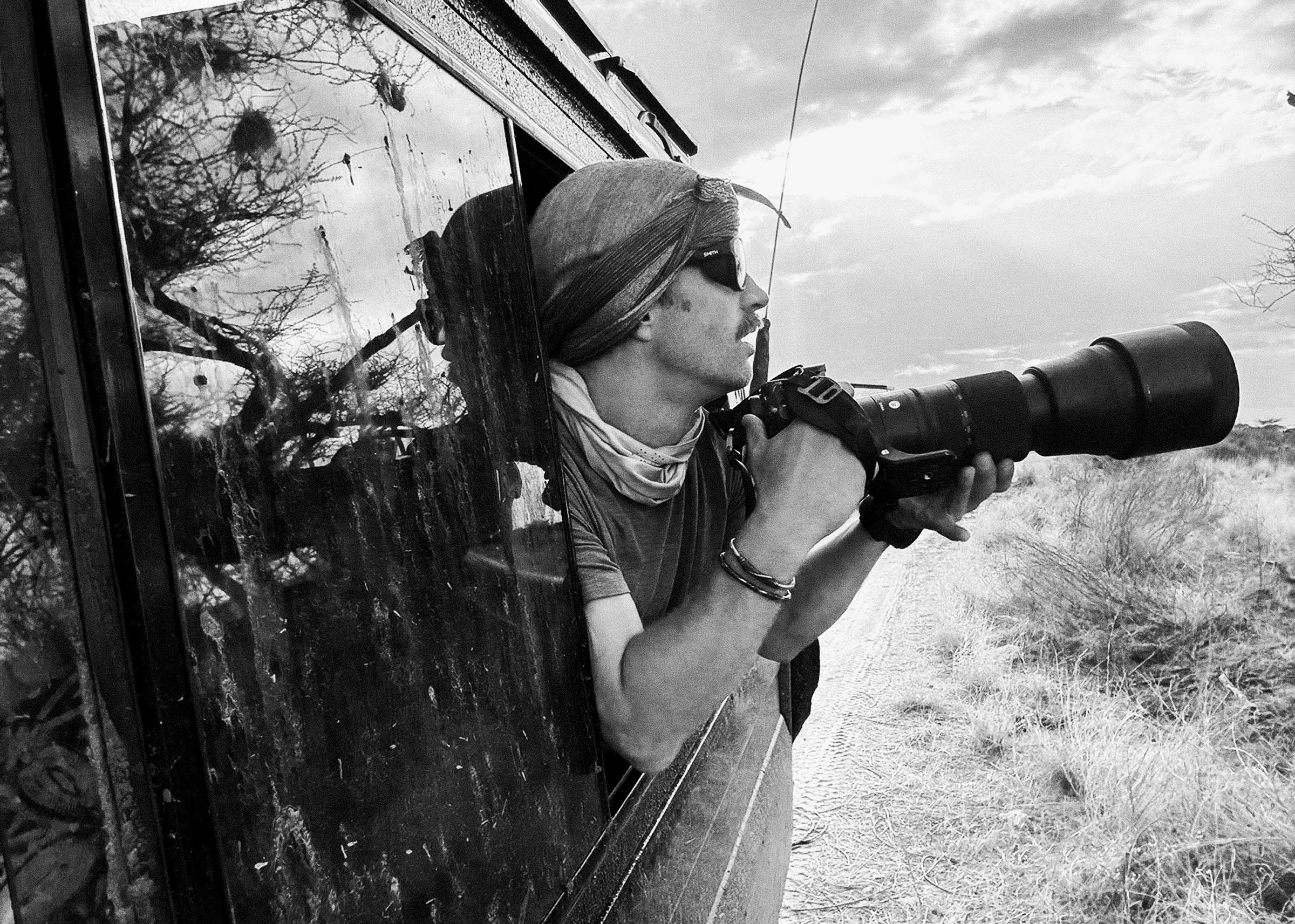 Zach Suggs wearing Smith sunglasses and a headscarf, looking through a camera with a long telephoto lens, leaning out of a vehicle window.