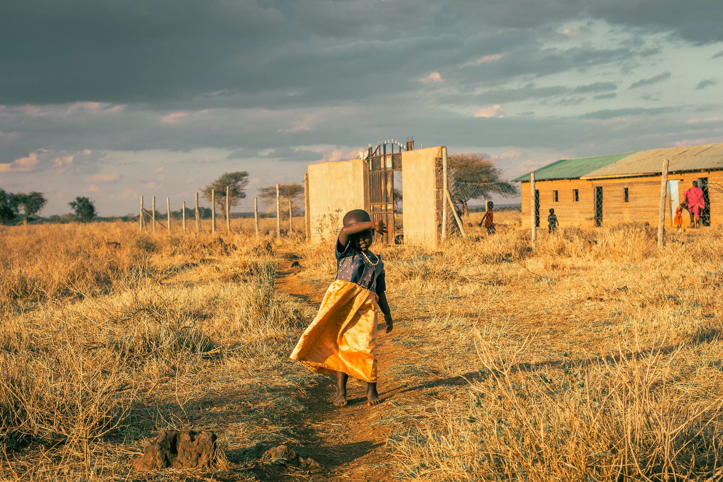 Young girl walking on a dirt path through dry grass in a rural area with a concrete gate, wooden house, and other children and adults in the background under a cloudy sky.