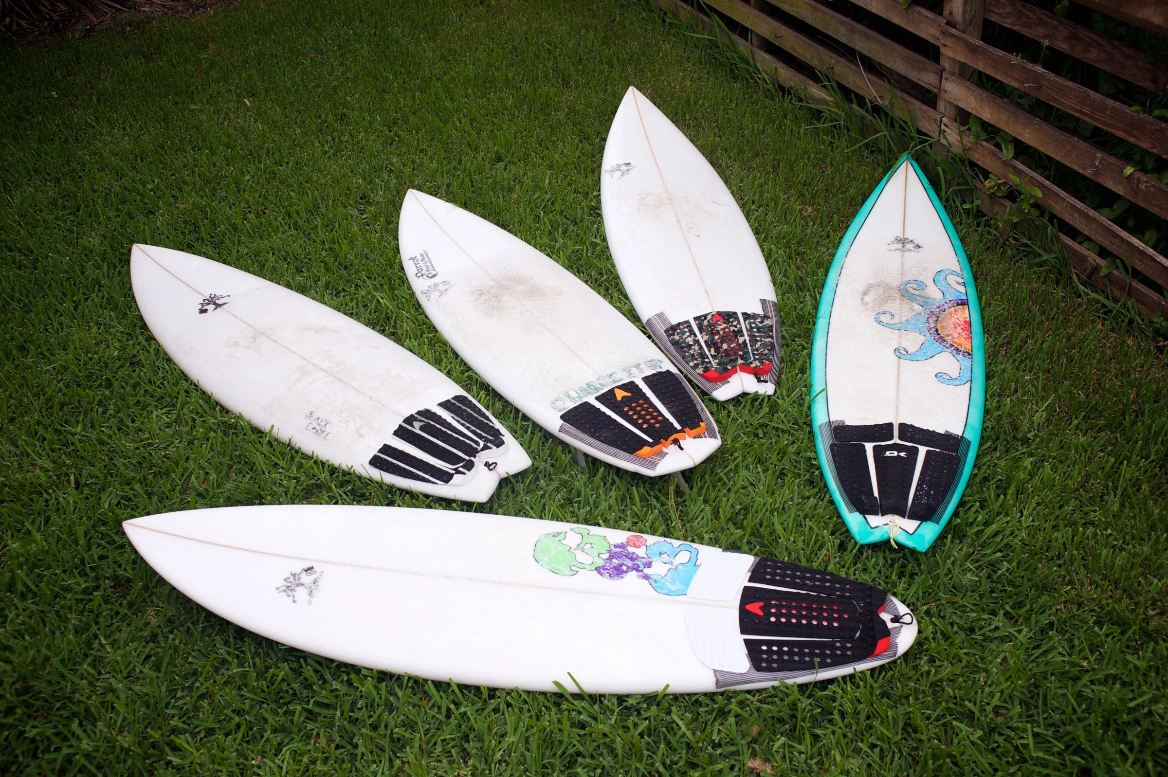 Five surfboards with colorful designs and traction pads, laid on green grass near a wooden fence.