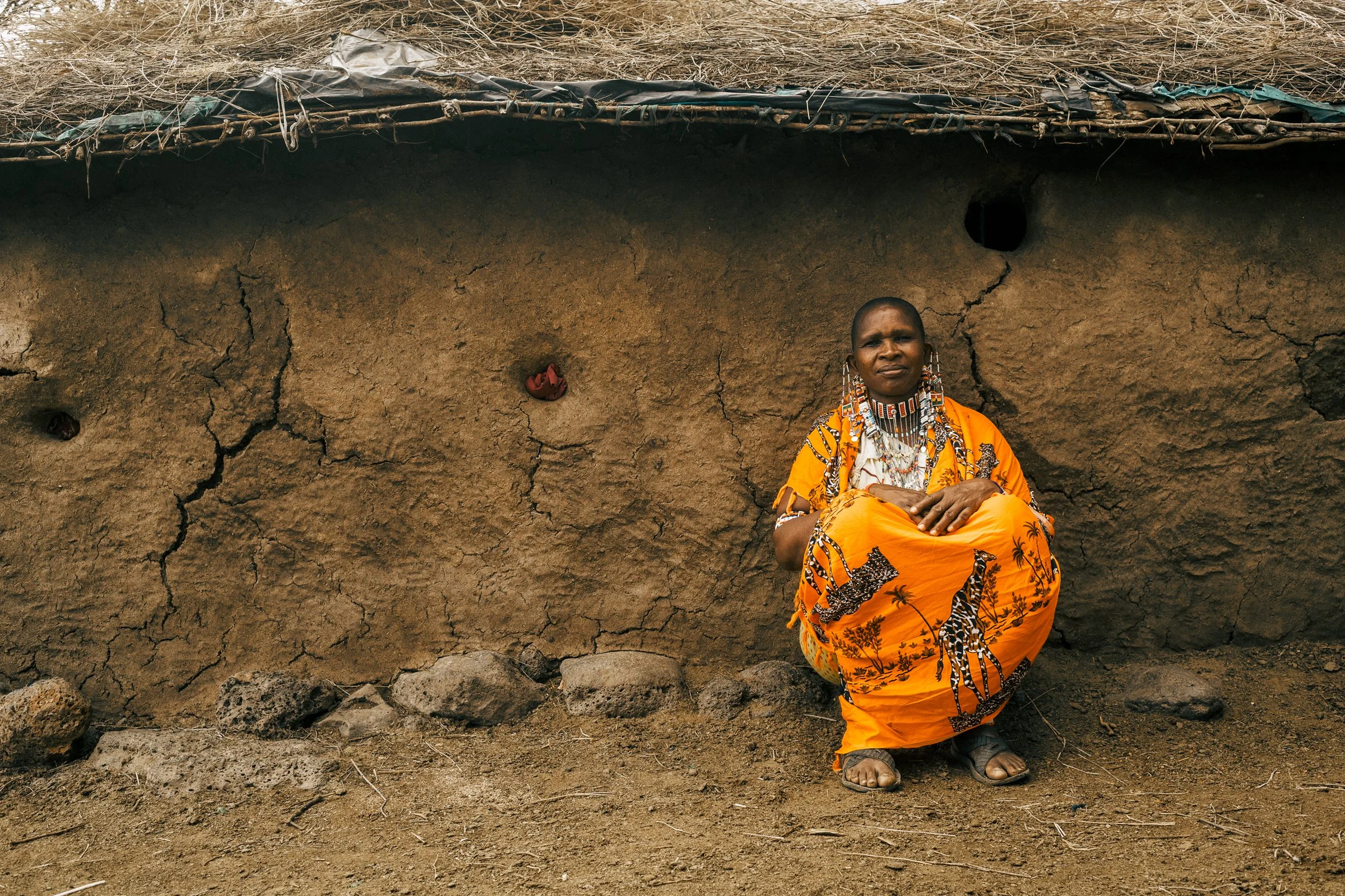 An indigenous woman in a bright orange traditional dress with beaded jewelry sitting against a large, cracked mud wall.