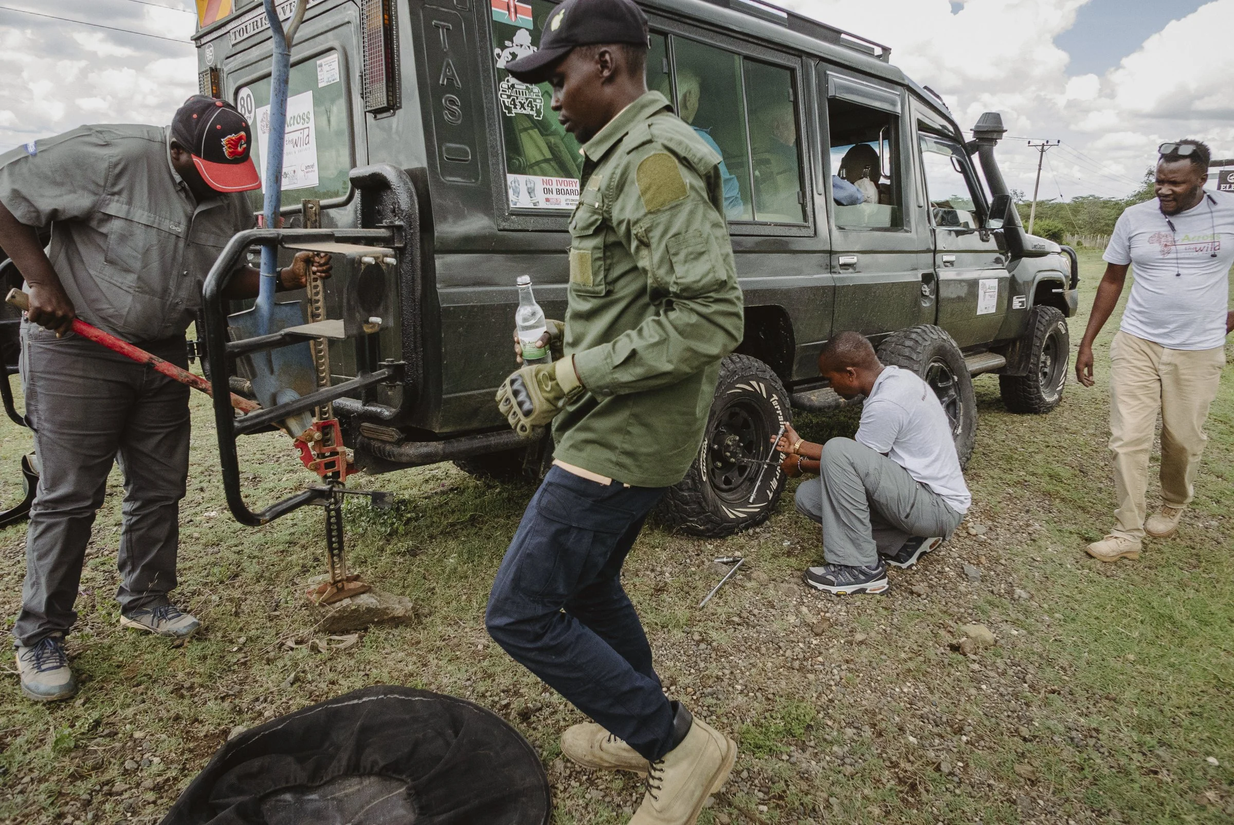 People working on a black off-road vehicle outdoors with cloudy sky and power lines in the background.
