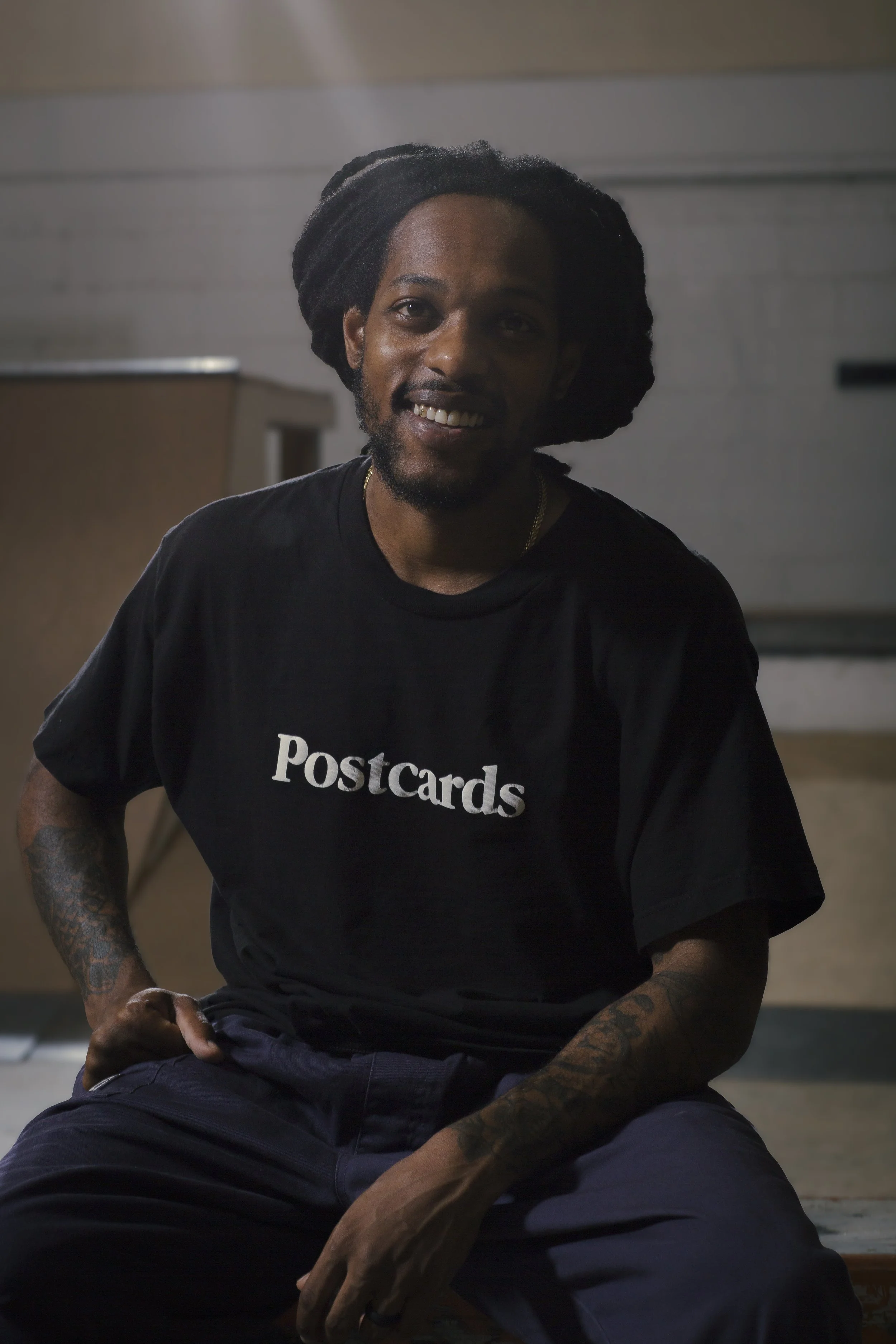 A man with dreadlocks, a beard, and tattoos on his arm, smiling and sitting on the ground wearing a black t-shirt that says 'Postcards'.