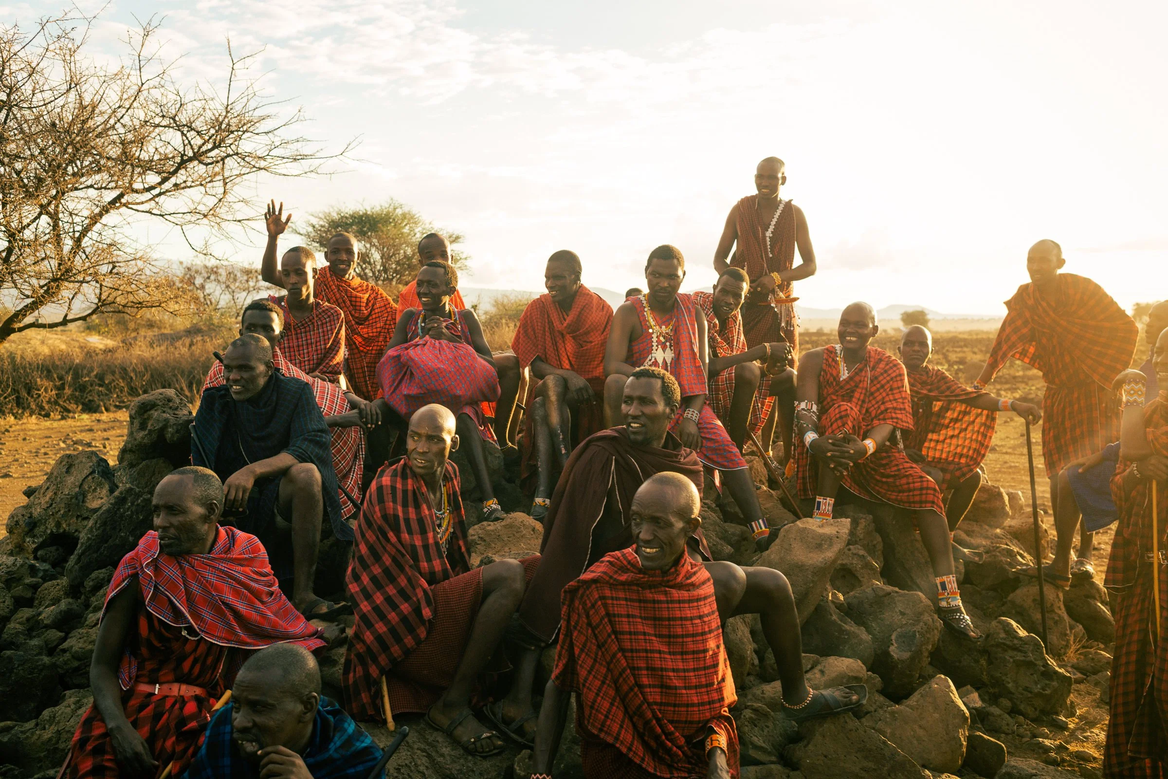 Group of Maasai people sitting and standing outdoors in a natural landscape during sunset, dressed in traditional red and patterned shukas, some smiling and engaging with the camera.