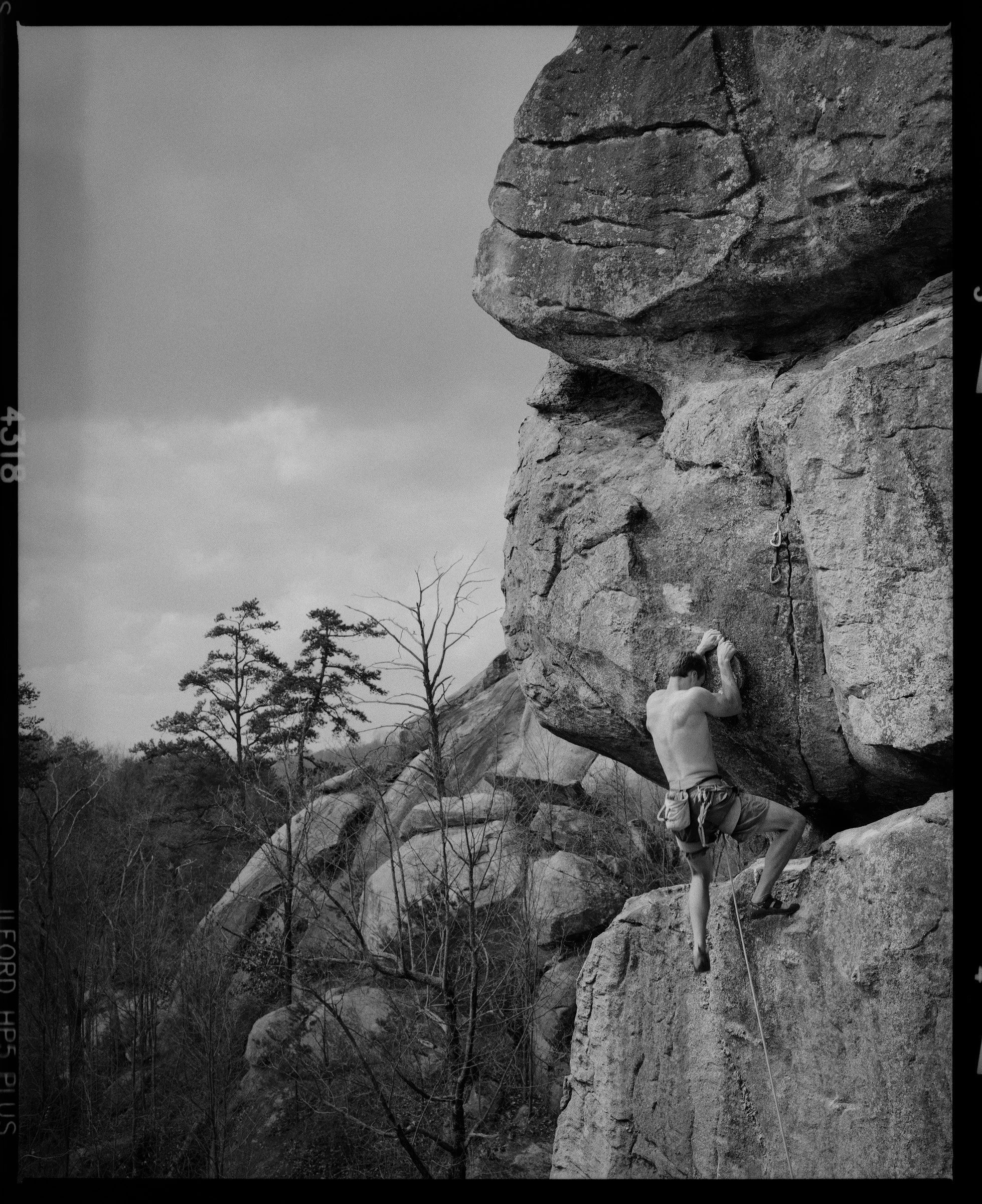 A person bouldering on a large outdoor rock formation surrounded by trees, in black and white.