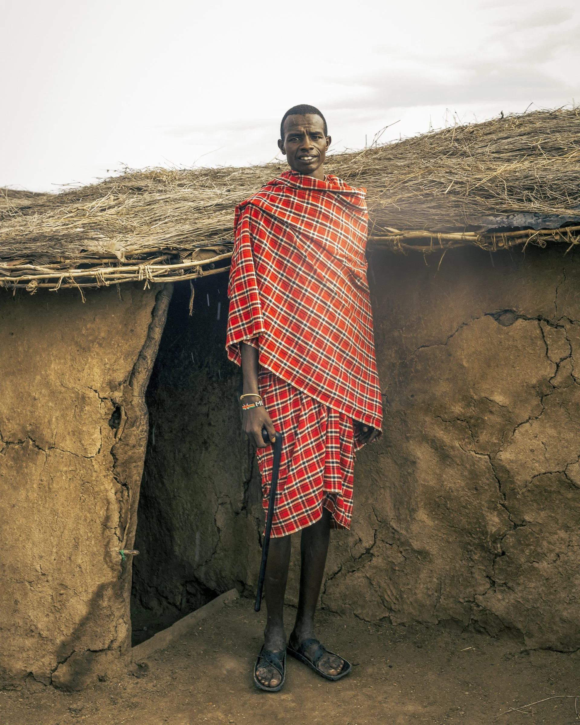 A man wearing a red plaid shawl and sandals standing outside of a traditional mud house with a thatched roof, holding a walking stick.