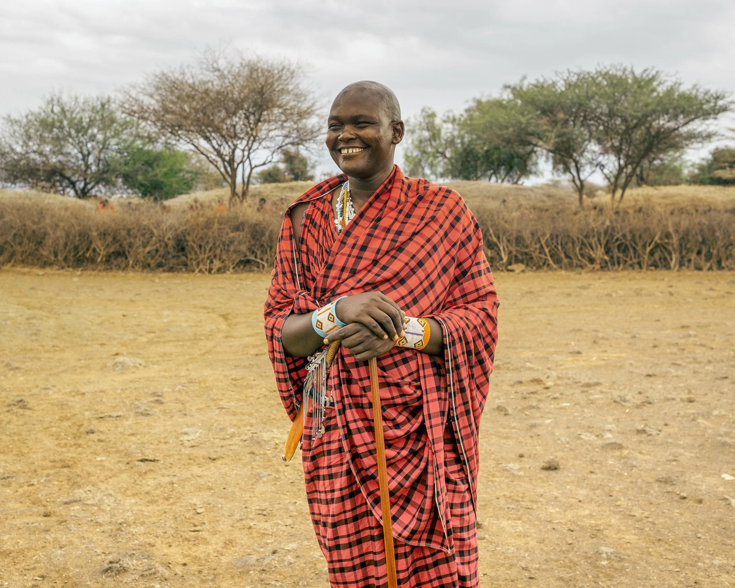 A smiling person with shaved hair wearing traditional Maasai clothing and jewelry, standing outdoors in a dry landscape with leafless and green trees in the background.