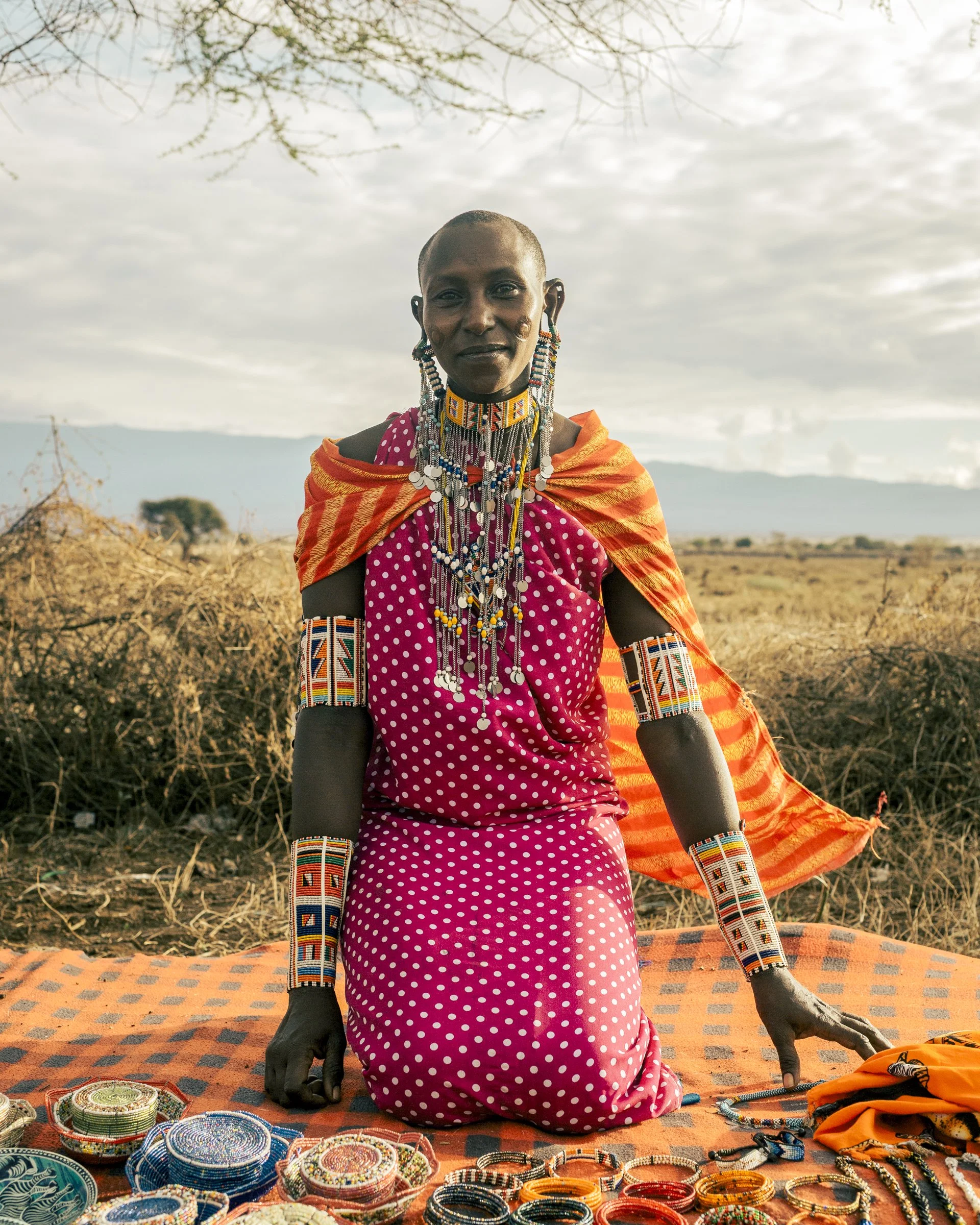 A woman in traditional Maasai attire sells beaded jewelry at an outdoor market in a rural area, with dry grass and a cloudy sky in the background.