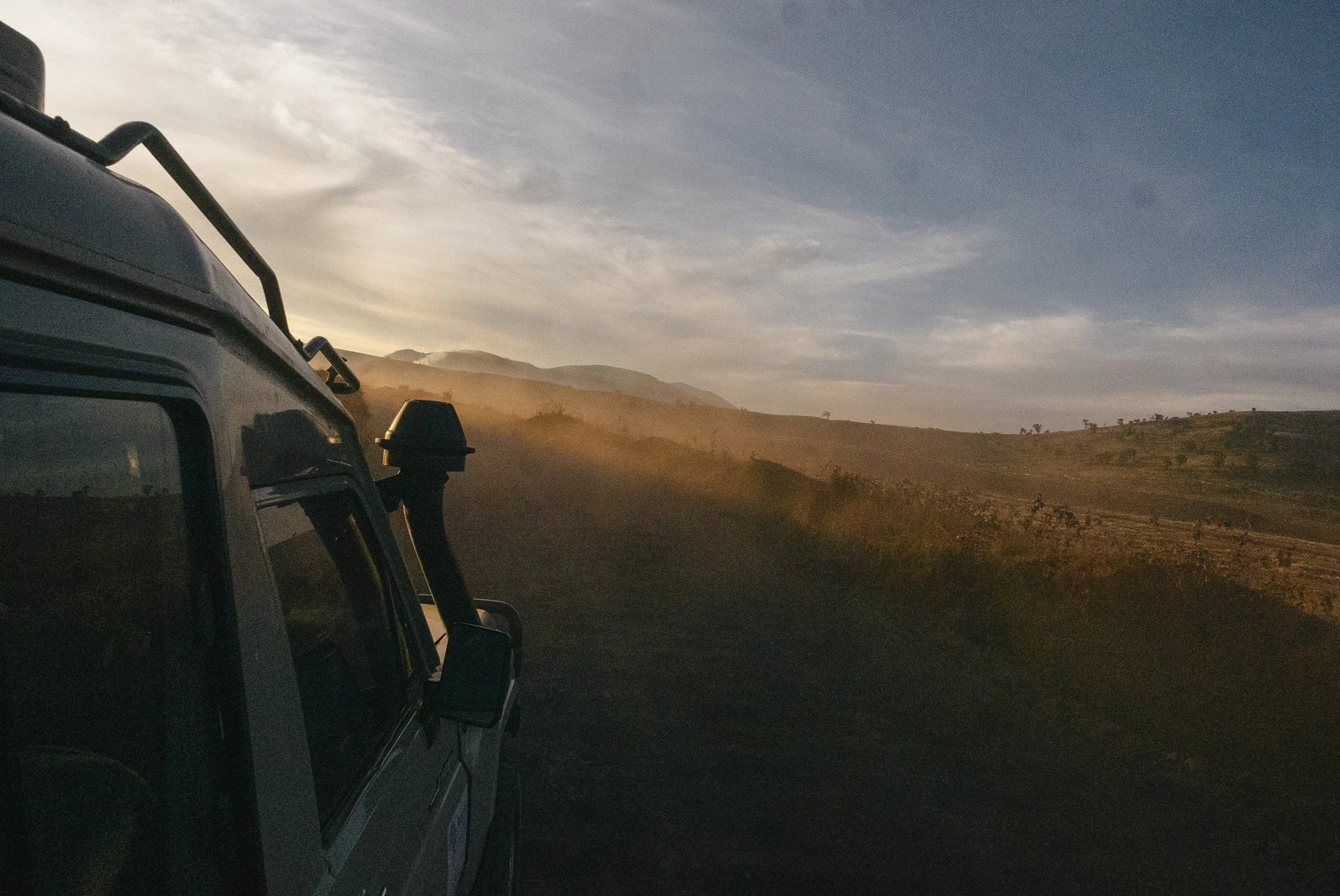 View from the side of a vehicle driving on a dusty road through a hilly landscape during sunset with a partly cloudy sky.