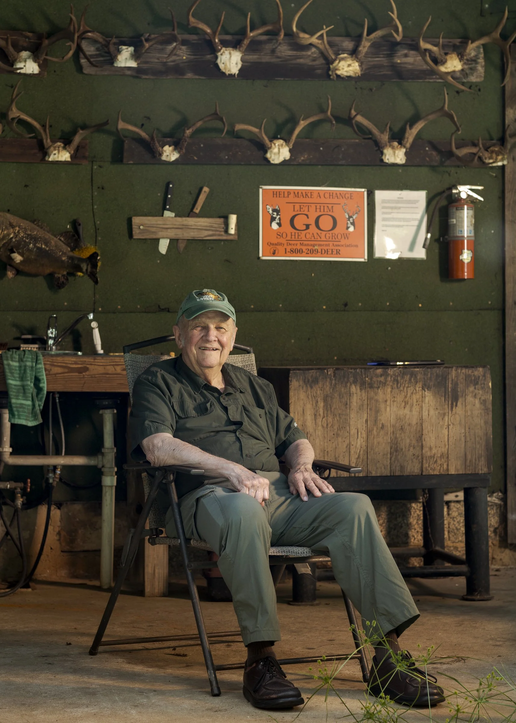 An elderly man sitting in a chair with a big smile, wearing a green cap, black shirt, and khaki pants in a rustic setting with deer antler decorations and a sign about deer management on the wall.